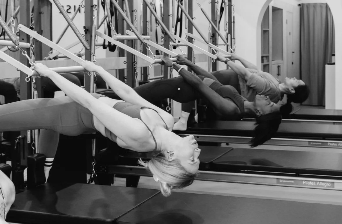 A woman is exercising on a reformer Pilates machine, using a pulley and spring attachment to perform a resistance exercise, with two children lying down on mats in the background.