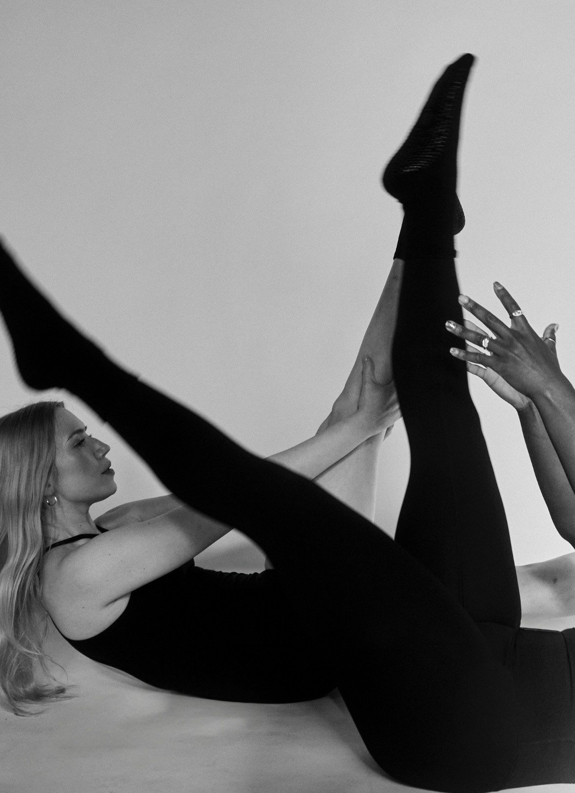 A woman with long blond hair performing a Pilates exercise on a black mat in a fitness studio.