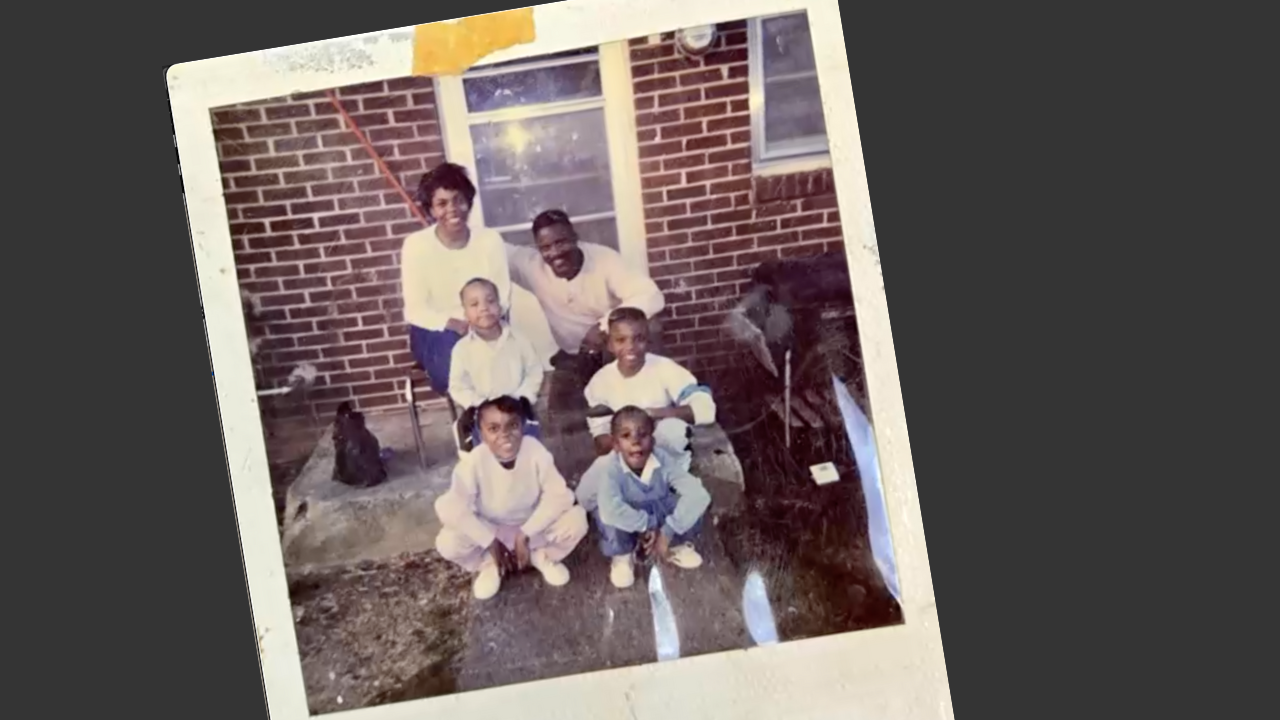 A family photo taken outside of a brick house featuring two adults and four children, with a dog and patio furniture.