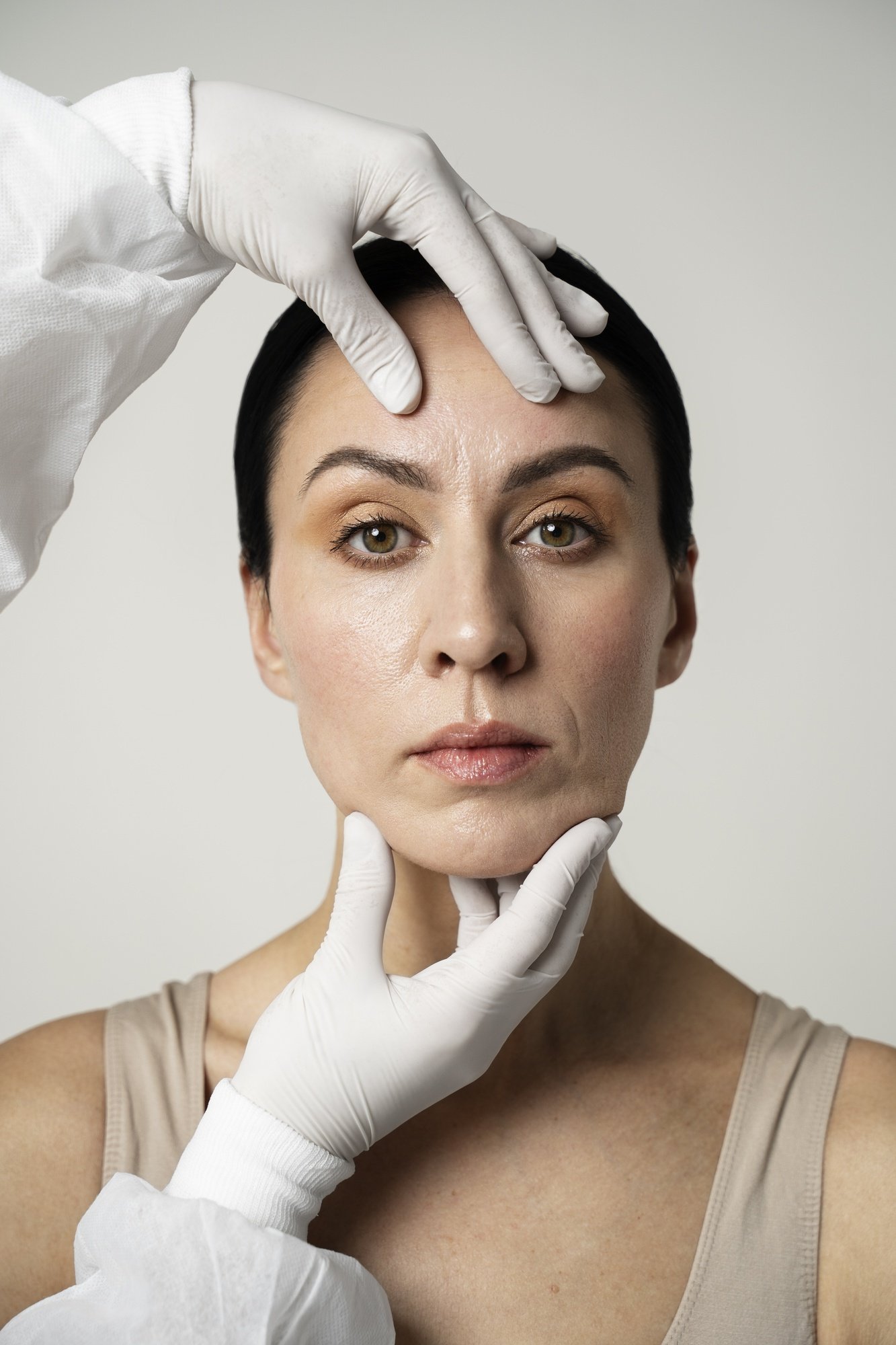 A person with dark hair is being examined by white-gloved hands on their face, indicating a medical or cosmetic evaluation.