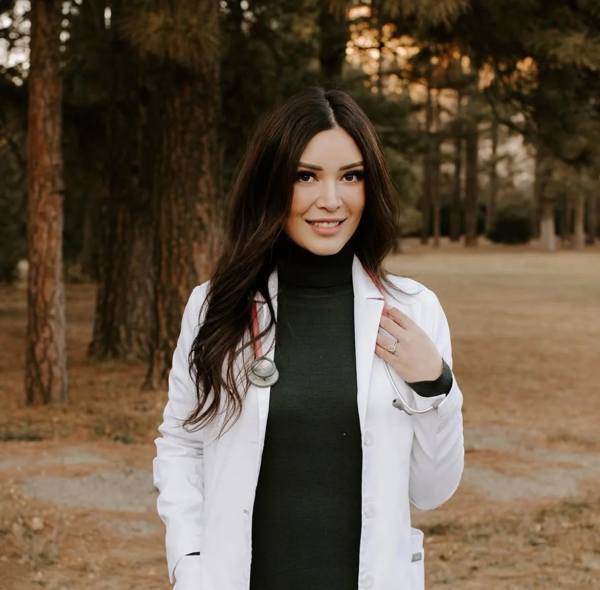 A woman in a white coat with a stethoscope standing outdoors in a wooded area.