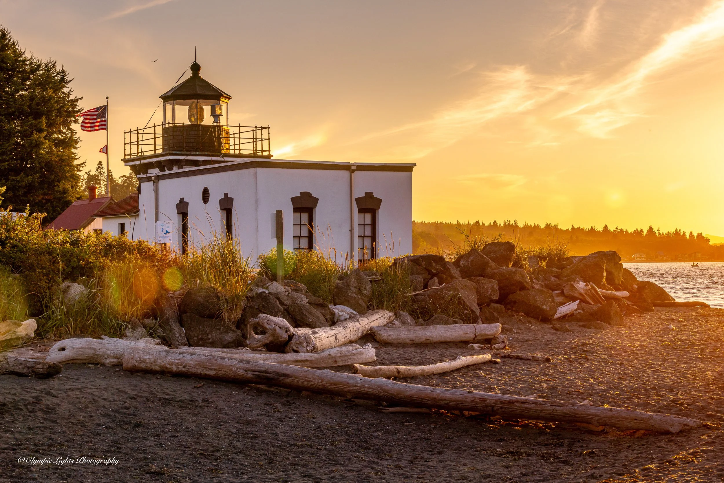 A lighthouse by the water during sunset with driftwood on the black sandy beach in the foreground, surrounded by rocks and vegetation.