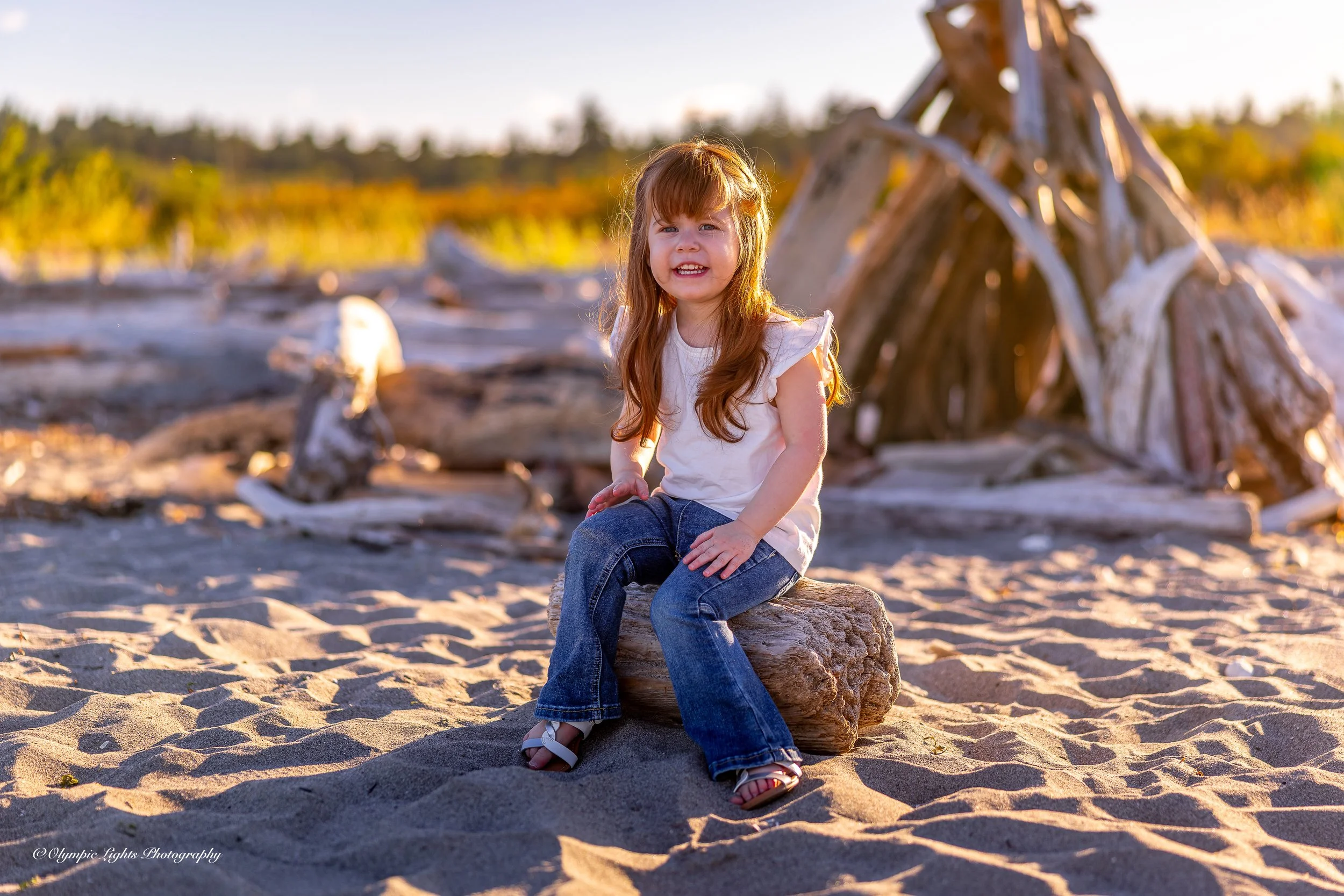 A young girl with long red hair wearing a white t-shirt and jeans sitting on a large piece of driftwood on a sandy beach during golden hour, with driftwood structure in the background.