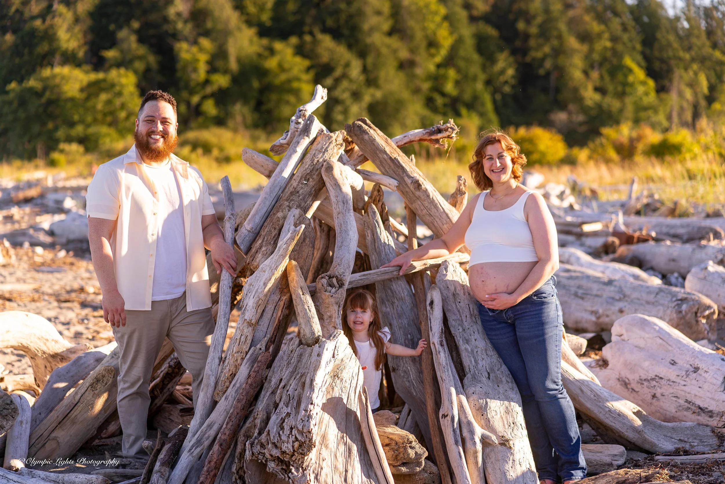A family of three, including a pregnant woman, on a beach with driftwood arranged in a teepee structure. They are smiling and enjoying the outdoor scenery with trees in the background.