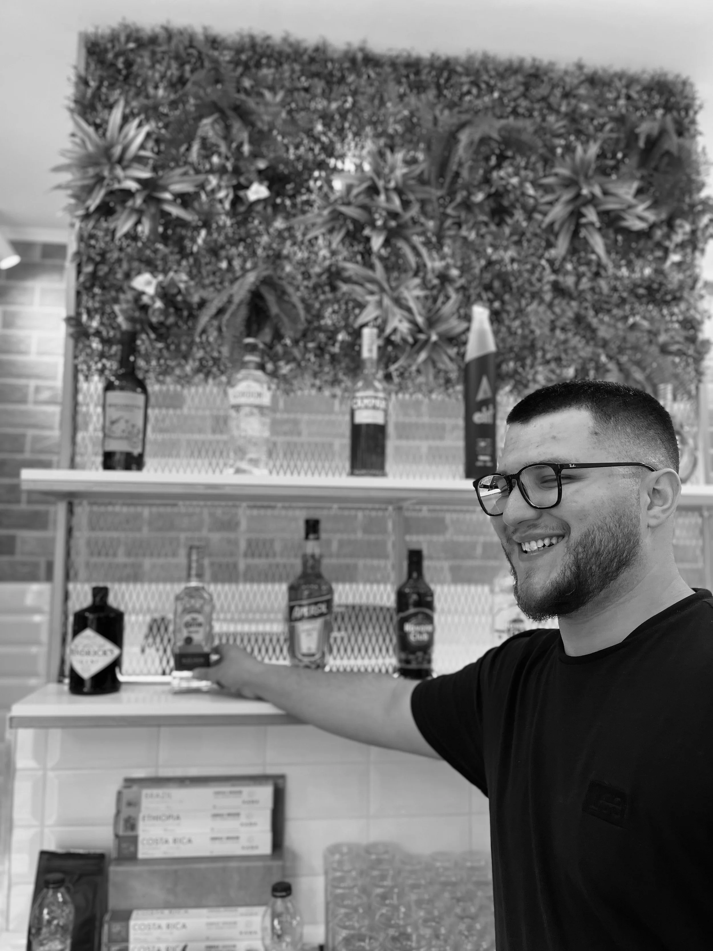 A smiling man with glasses extends his arm towards liquor bottles displayed on a shelf, with a decorative leafy background behind him.