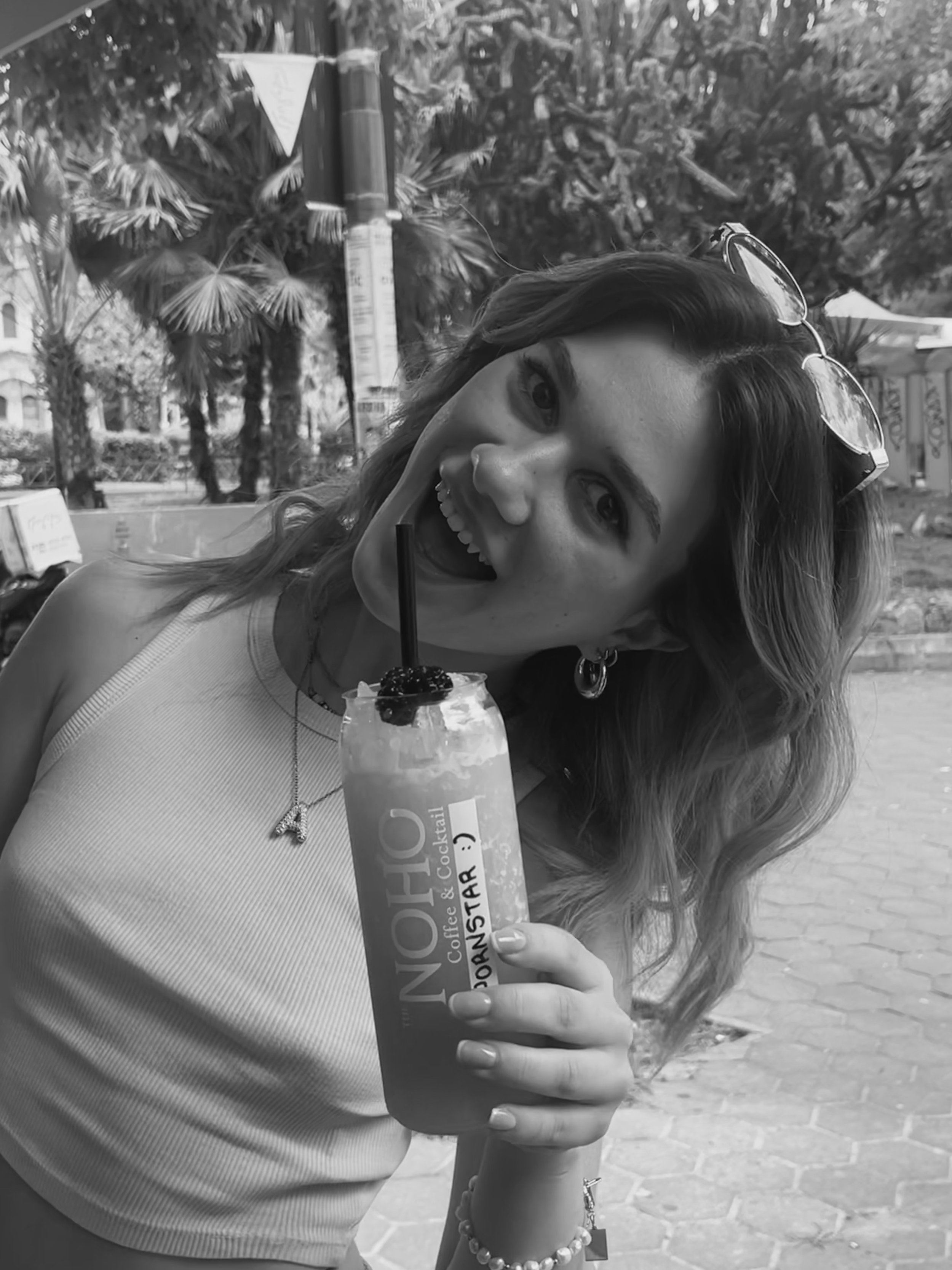 A woman with wavy hair wearing sunglasses on her head, smiling with an open mouth, holding a milkshake or smoothie with a straw, garnished with berries, outdoors on a sunny day.