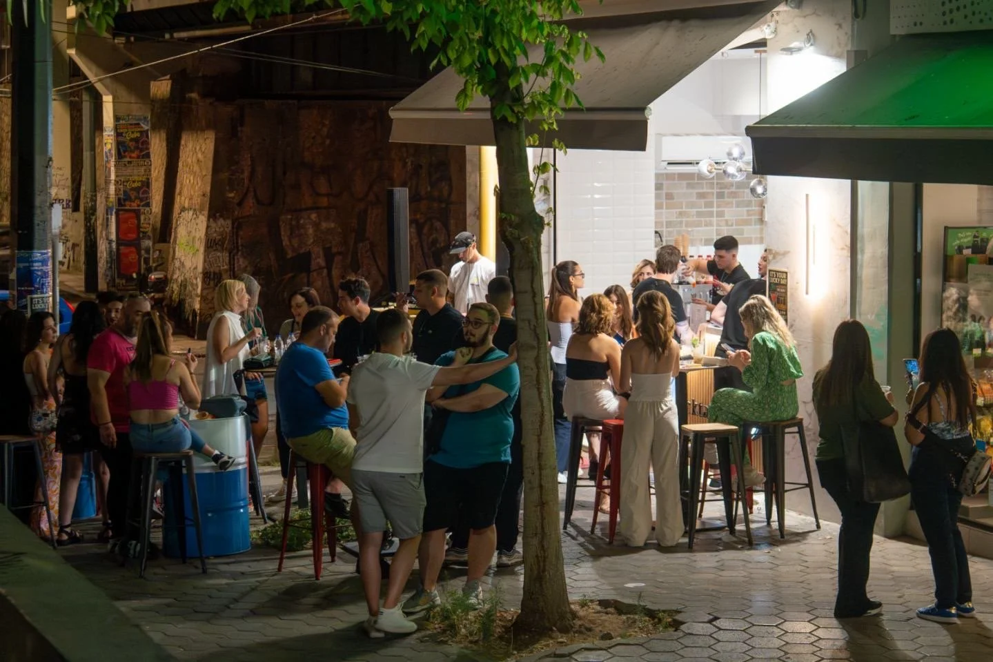 People gathering at an outdoor bar or food stand during the evening, with some ordering and others socializing under green and white umbrellas.
