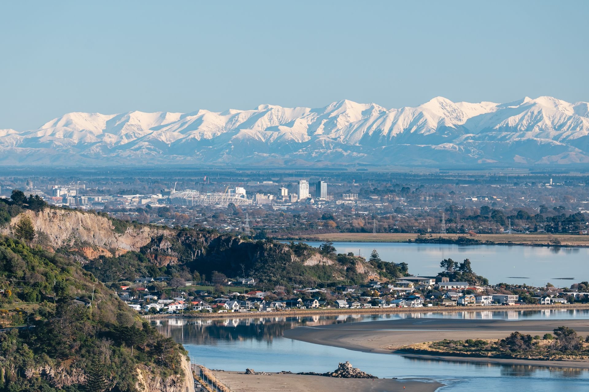 Snow-capped mountains in the background, cityscape with buildings, river, and residential area in the foreground.