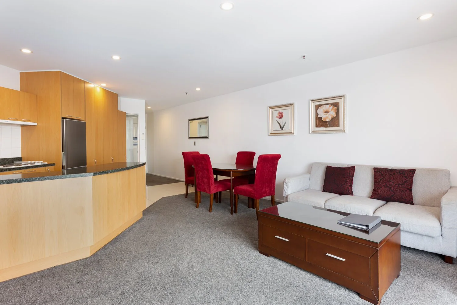 Modern open-plan living room with a kitchen and dining area. The room features a gray sofa with red cushions, a wooden coffee table, and a round dining table with red chairs. The kitchen has wooden cabinetry and a dark countertop. Two floral painting