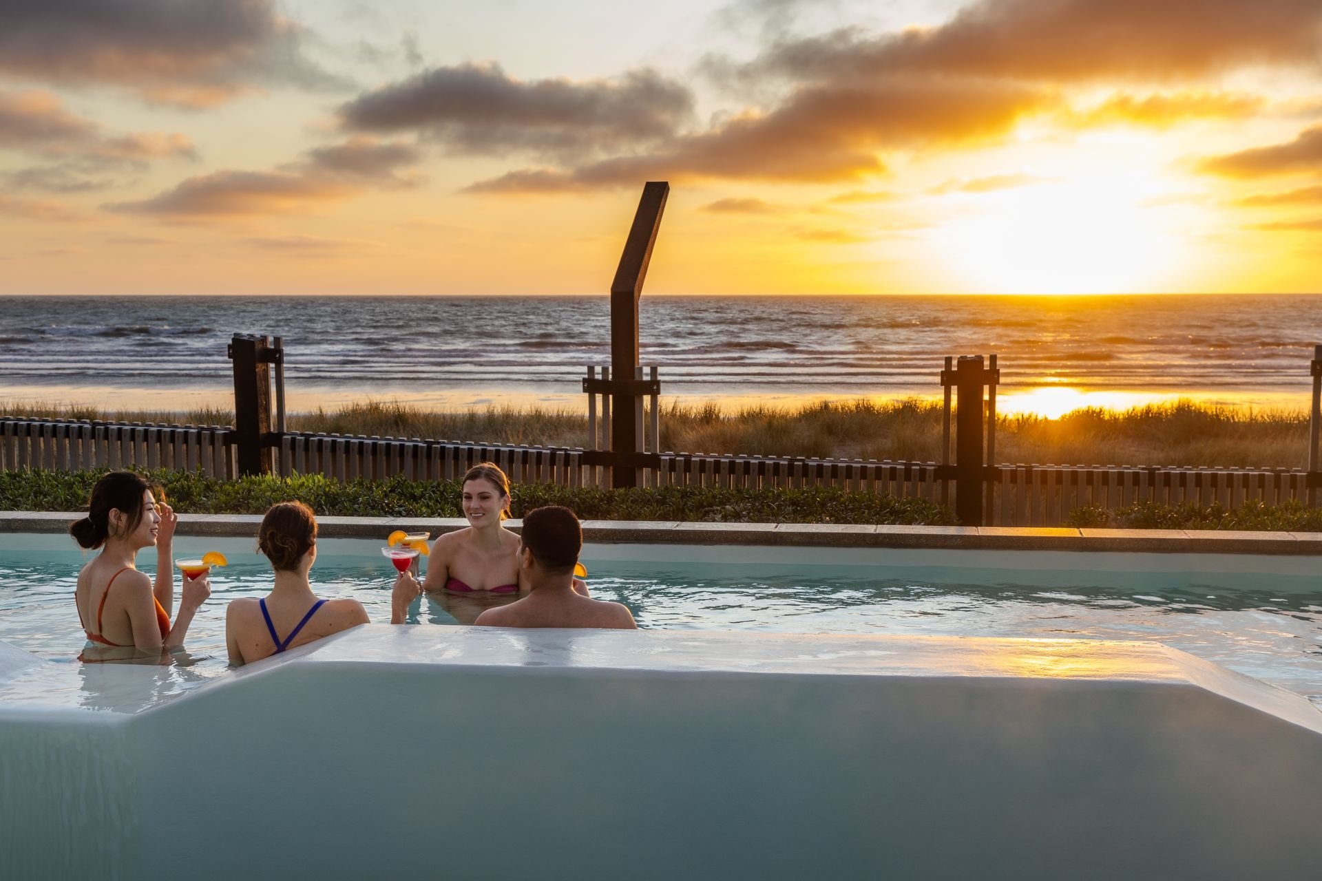 Four people in a pool with drinks at sunset, overlooking the ocean.