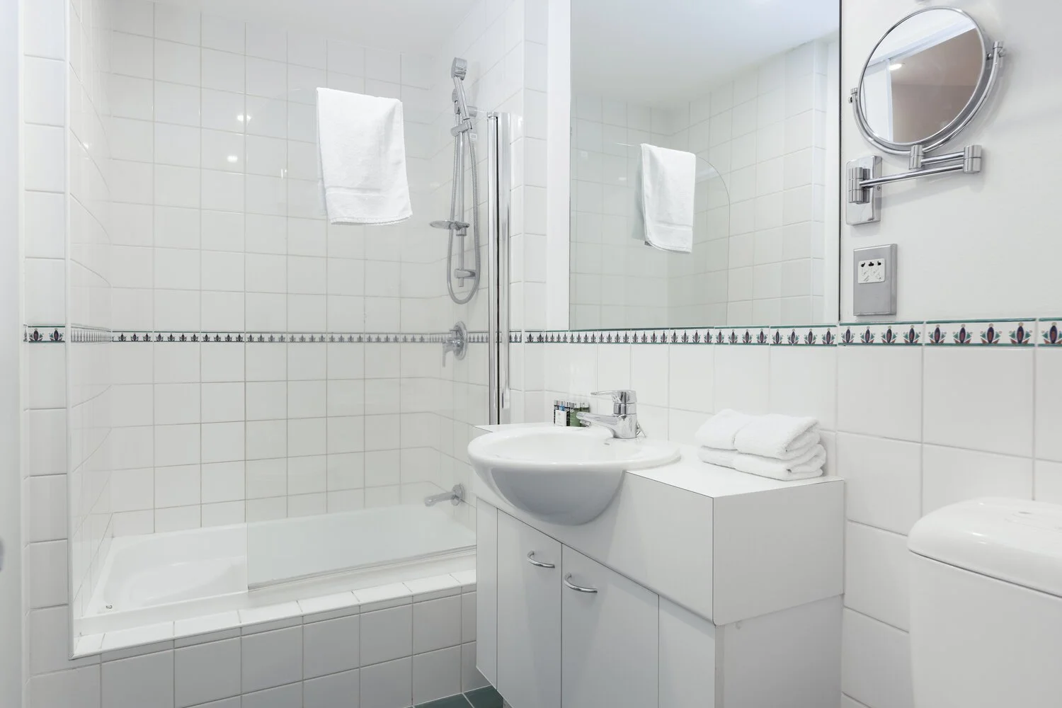 Modern white bathroom with tiled walls, a bathtub with a shower, wall-mounted towel rack, sink with a mirror, folded towels, and a toilet.