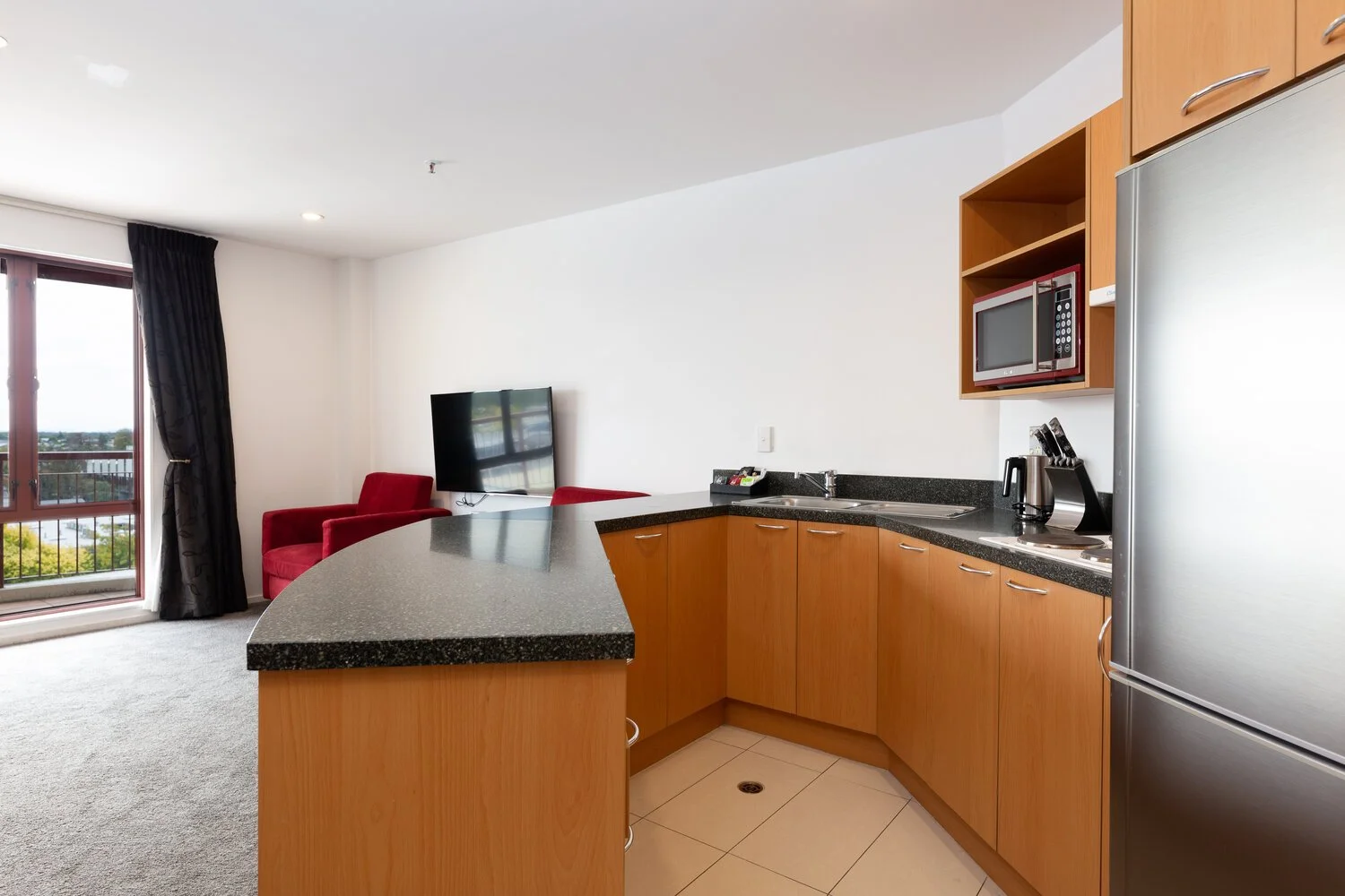 Modern kitchenette with wooden cabinets, stainless steel refrigerator, microwave, and sink. Living area features red chairs, a flat-screen TV, and large windows with a view.