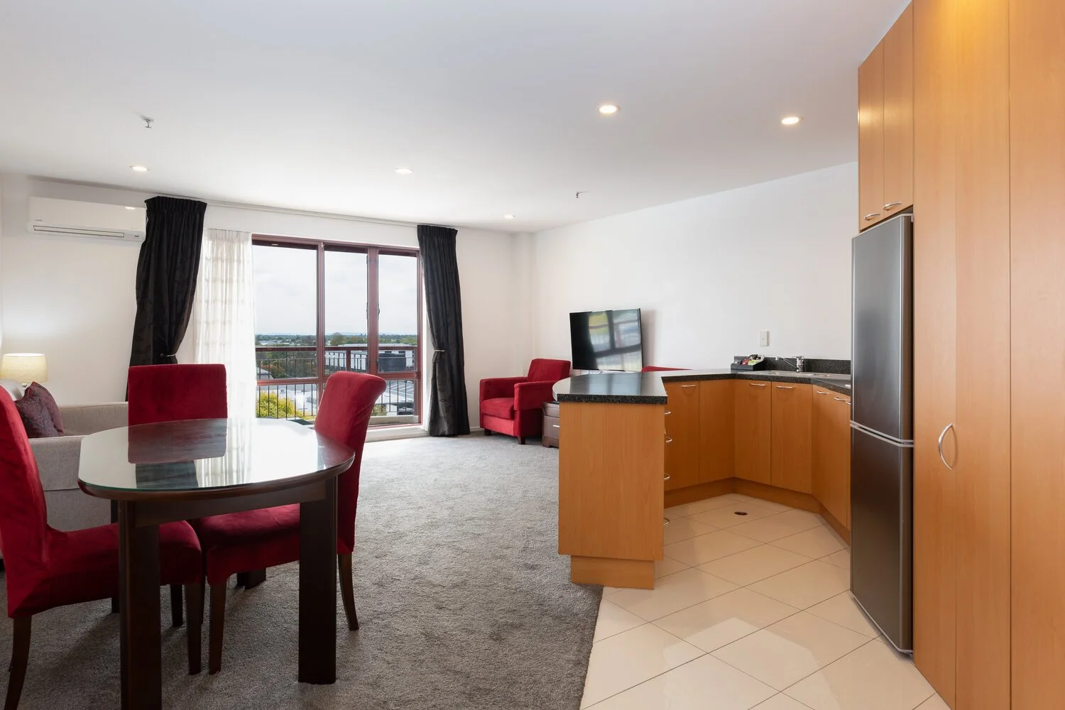 Modern open-plan living area with kitchenette, dining table with red chairs, armchair, TV, and large window with curtains.