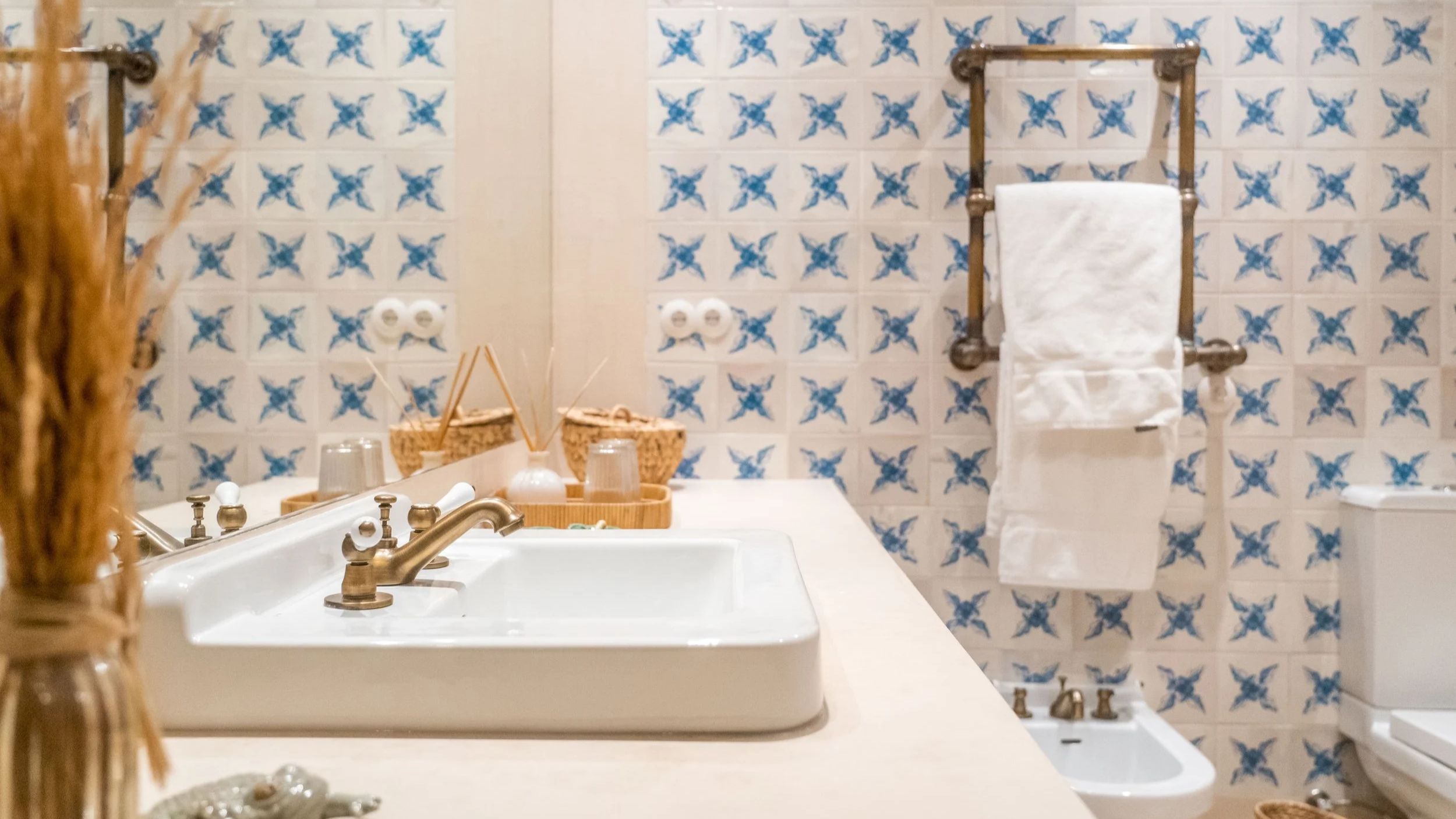 Bathroom with a white sink, brass fixtures, a basket with toiletries, a mirror, wall tiles with blue butterfly patterns, a towel rack with a white towel, a toilet, and a bidet.