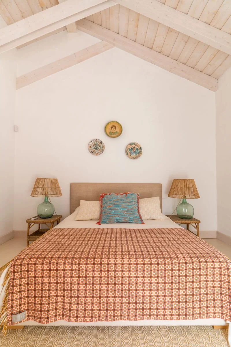 A bedroom with a bed, two bedside tables with lamps, and wall art overhead, featuring a white wall, a wooden bed frame, and a patterned orange coverlet.