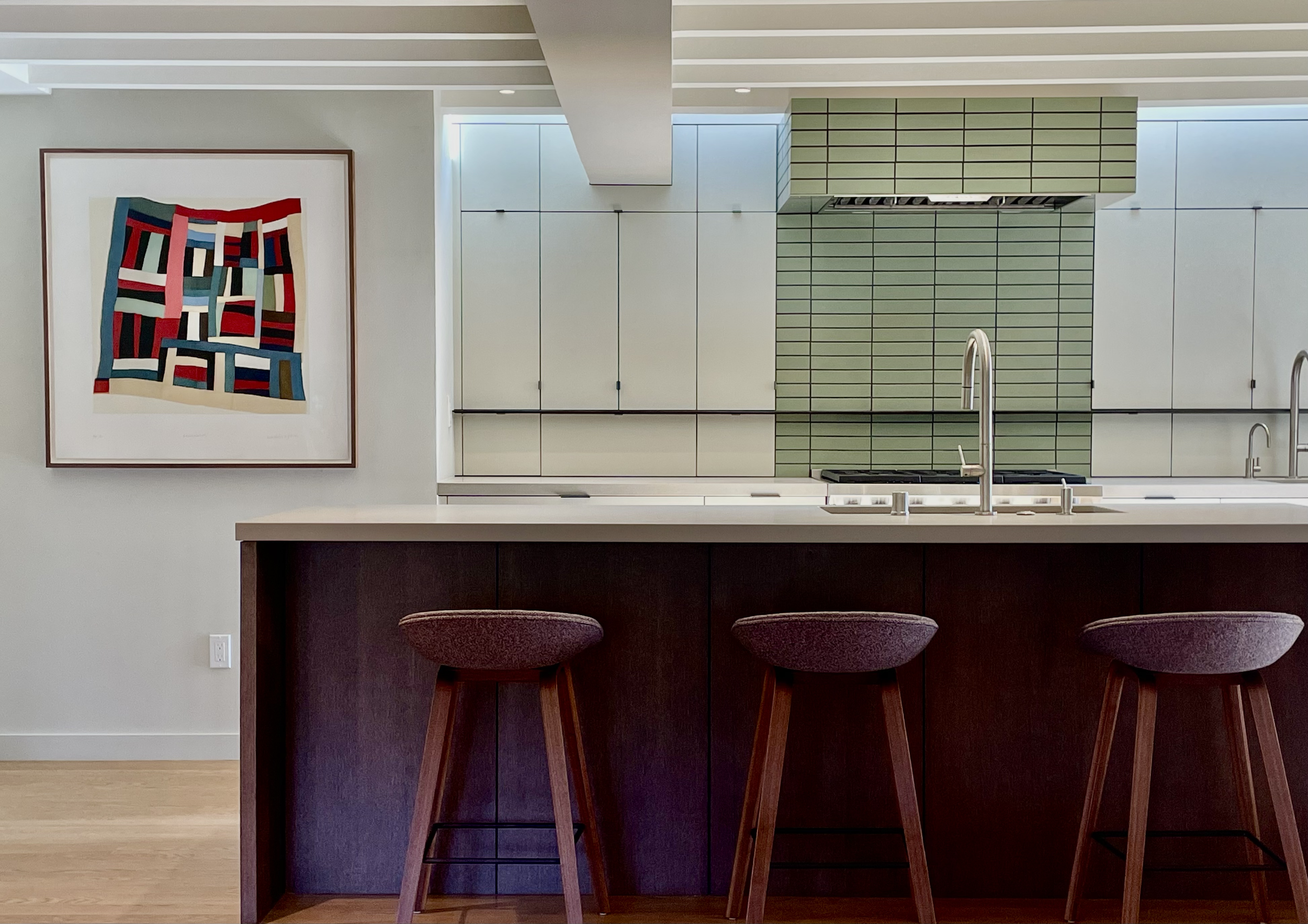 Modern kitchen with a white countertop island, three purple barstools, a green tiled backsplash, and a colorful abstract painting on a light gray wall.