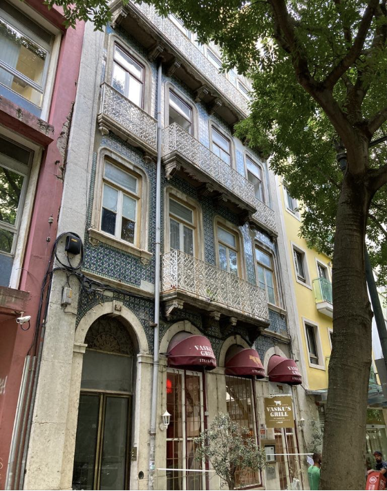 Historic building with ornate balconies and blue decorative tiles located on a busy city street, with a large tree in the foreground.