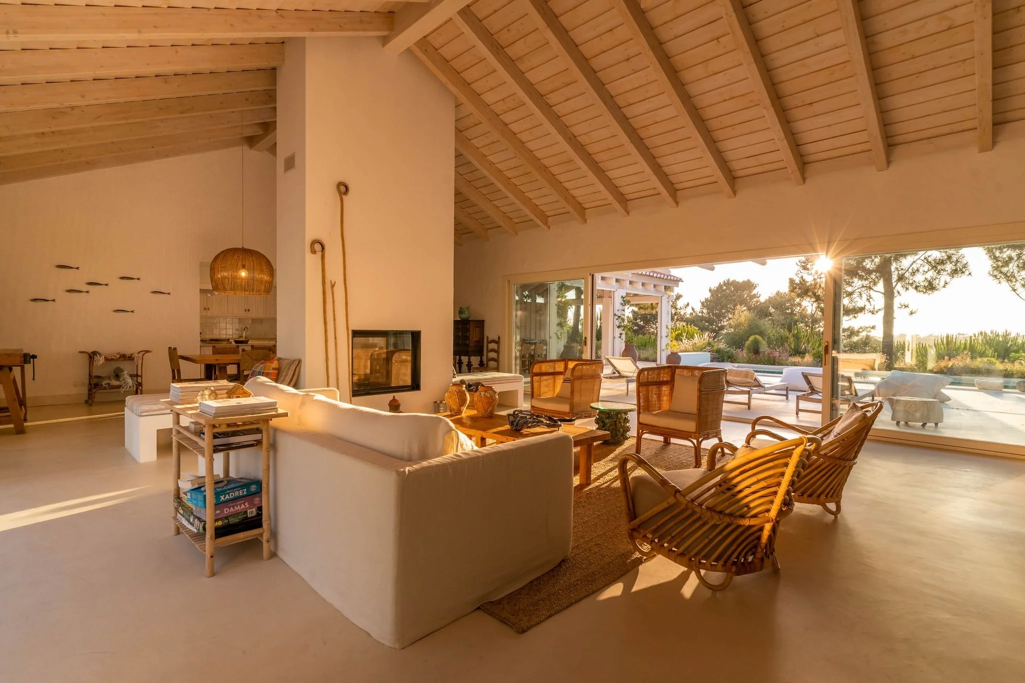 Bright living room with high wooden ceiling, large windows, and outdoor view, furnished with a white sofa, wicker chairs, and coffee table, decorated with pottery and lanterns.