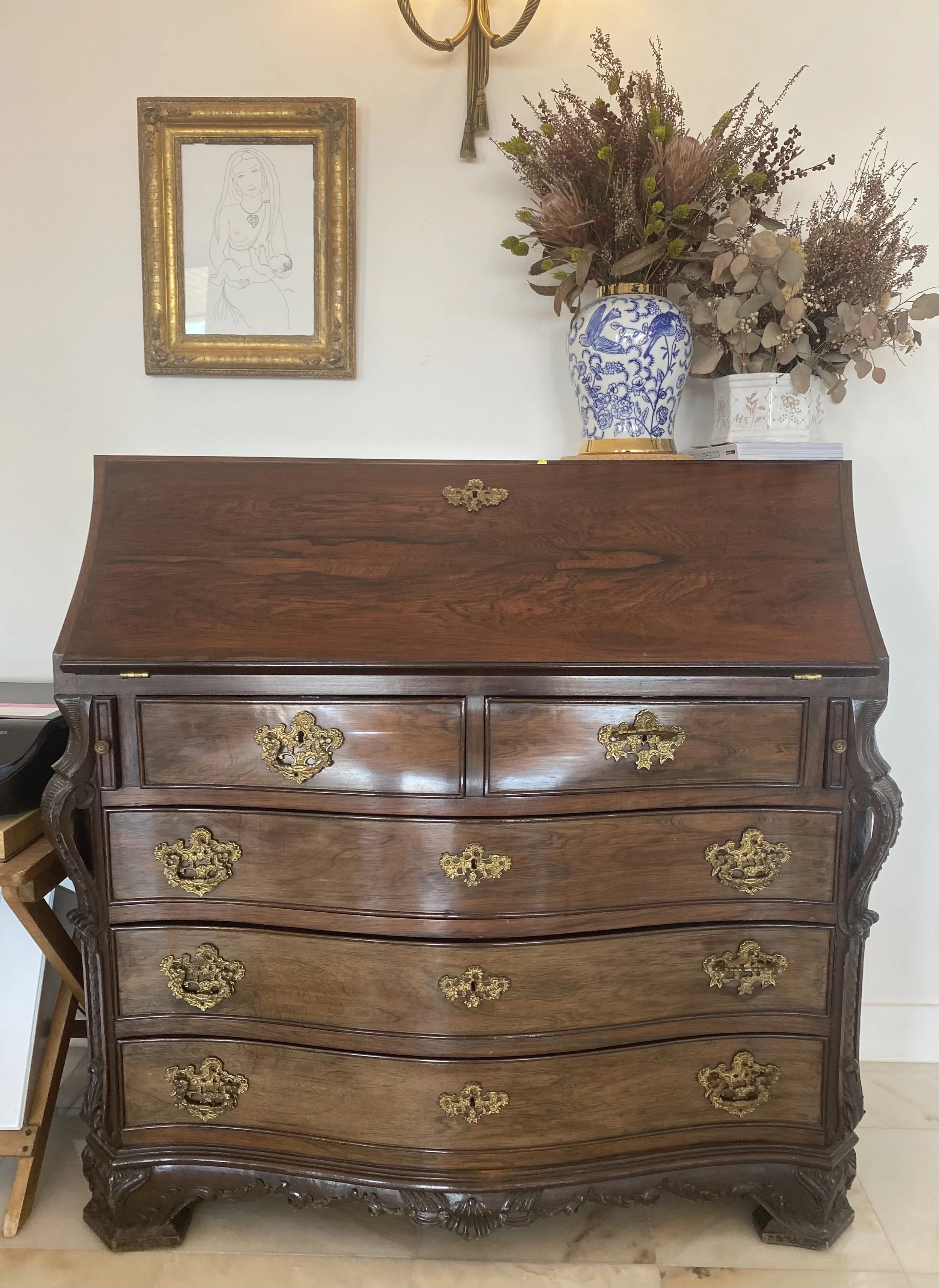 A vintage wooden dresser with ornate gold handles, decorated with brass accents, topped with a large floral bouquet in a blue and white vase, hanging on a white wall with a framed pencil drawing of a woman and a brass wall sconce above.