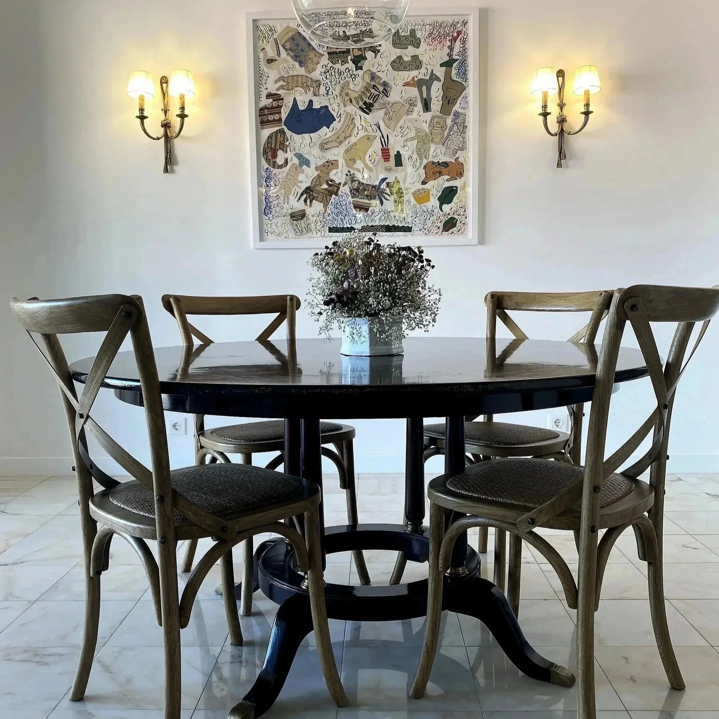 A round wooden dining table with five wooden chairs arranged around it. In the center of the table is a vase with white flowers. On the wall behind, there is a framed piece of abstract artwork with various shapes and colors. Two wall sconces with yellow lampshades are mounted on either side of the artwork.