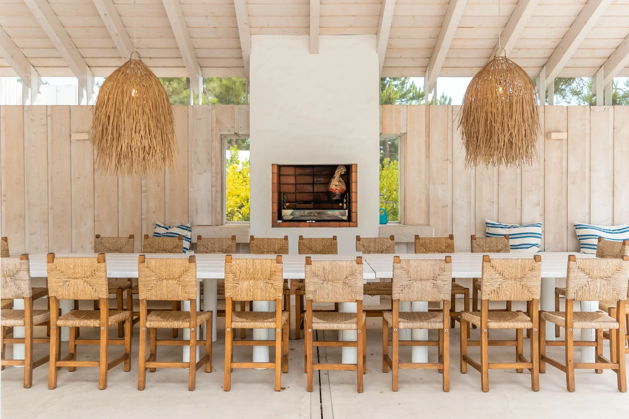 Interior of a light wood dining area with a long white table, wooden woven chairs, two hanging wicker light fixtures, a central white chimney with a built-in fireplace, windows showing greenery outside, and cushions on the bench seating.