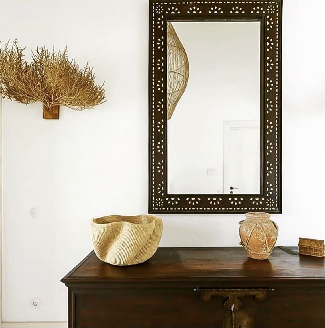 Decorative interior with a large mirror with a carved dark frame, a woven basket, a terracotta pot, a small wooden box, a wall-mounted art piece resembling a dried plant or tree, and a reflected paper lantern lamp.