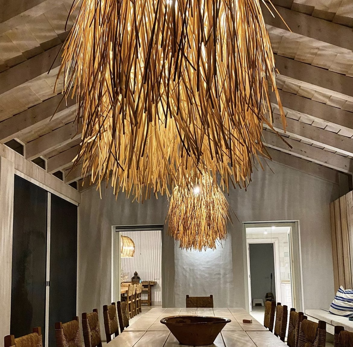 A dining room with large, hanging, straw-like pendant lights over a wooden table surrounded by chairs, with a view into a kitchen through an open doorway.