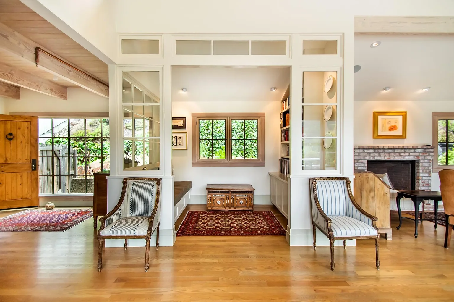 View of a cozy living space with two striped armchairs, hardwood floors, and a brick fireplace, featuring large windows and a mix of natural wood and white made interior design elements.