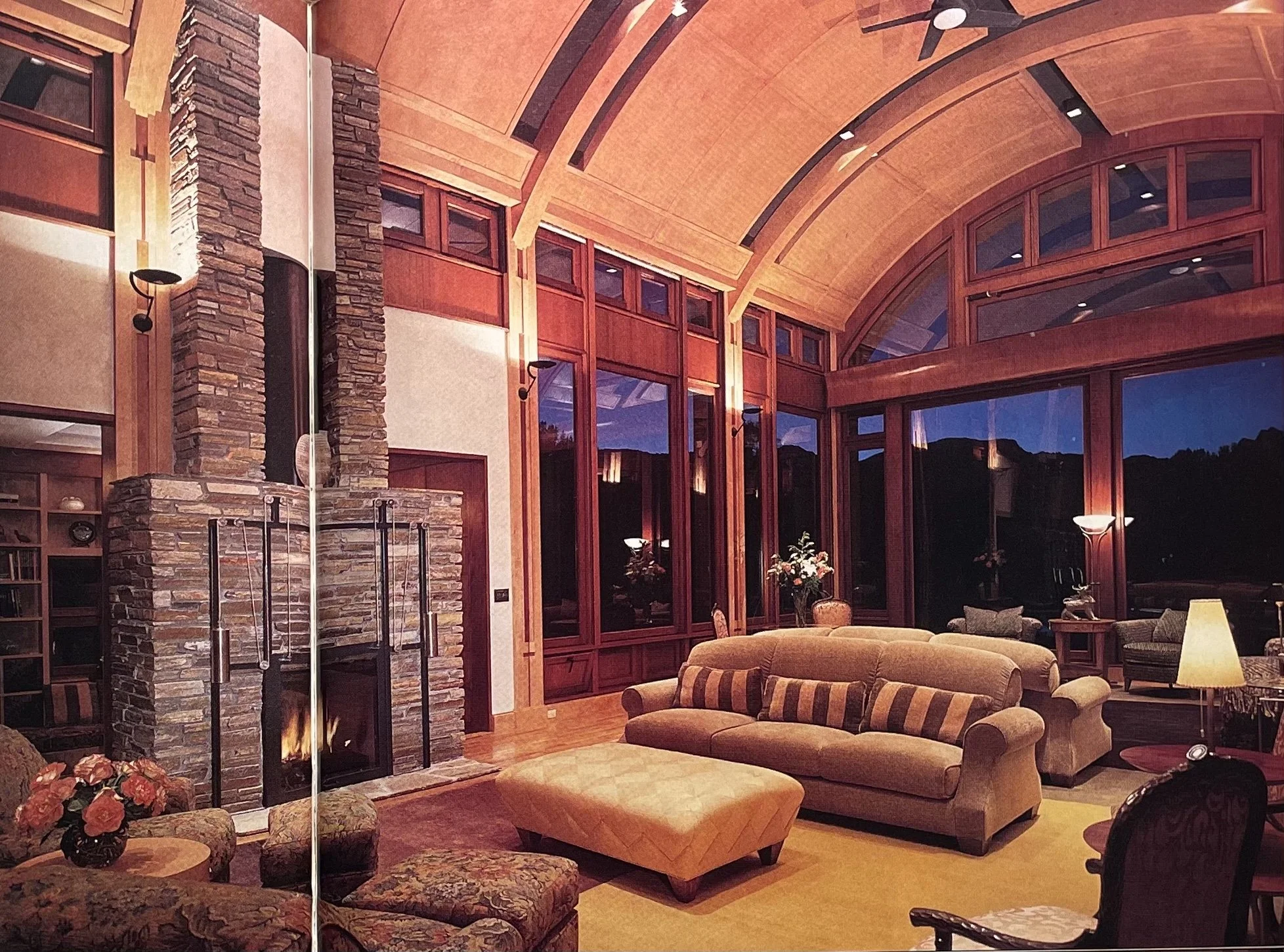 Living room with wooden vaulted ceiling, large windows, stone fireplace, beige sofas, and various chairs and lamps, evening scene