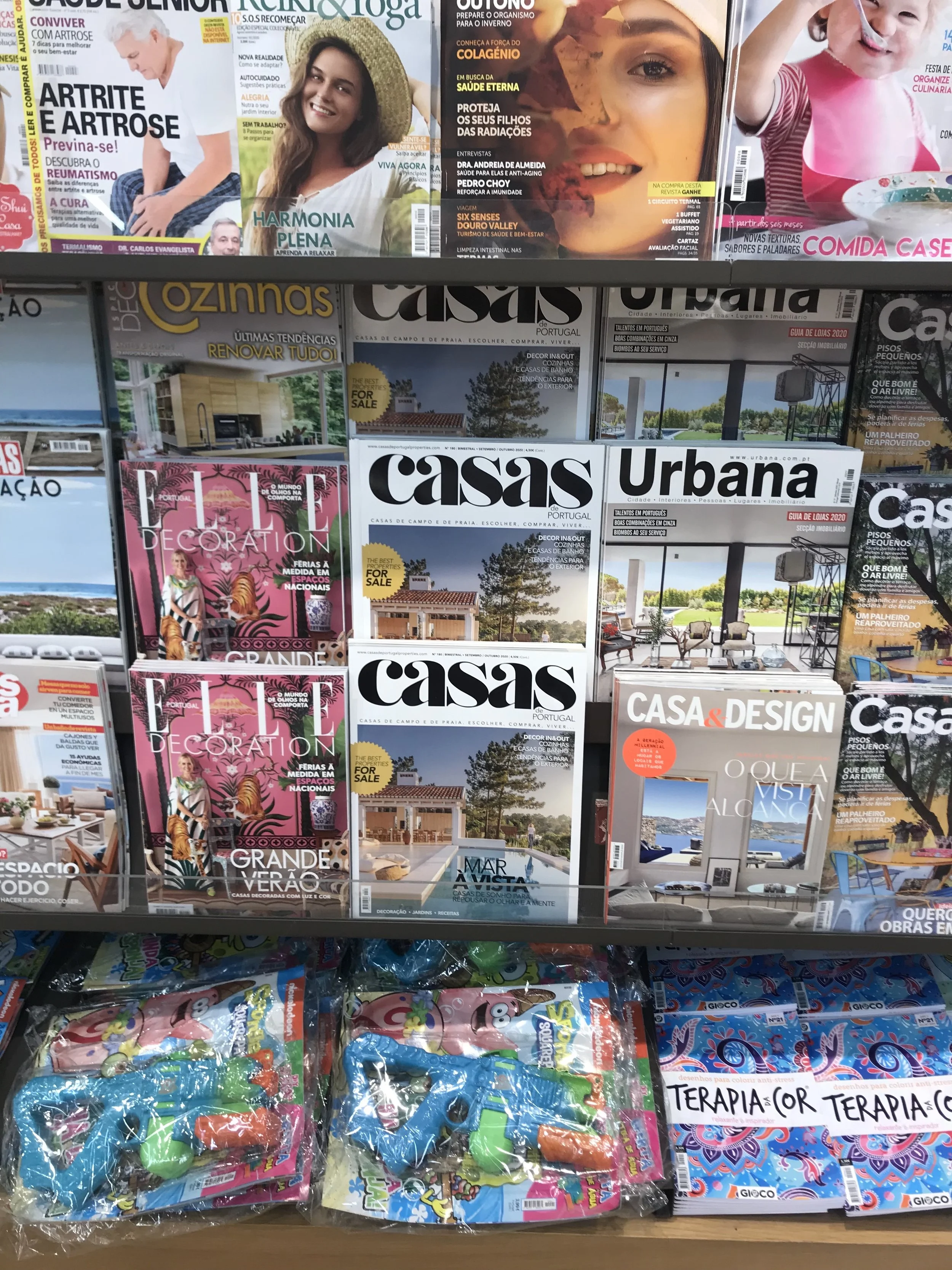 A display of magazines on a store shelf, including titles like 'Casa', 'Elle Decoration', and 'Casa & Design', as well as children's toys at the bottom.