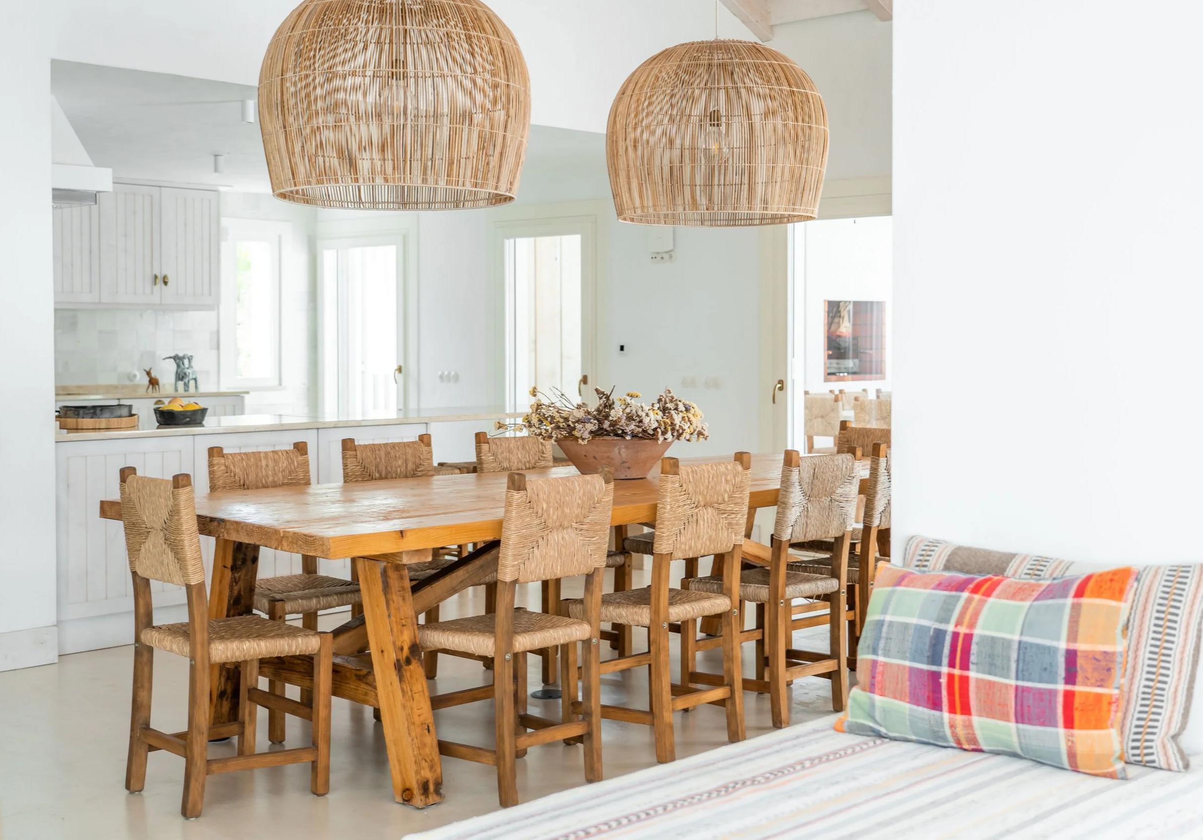 A rustic wooden dining table with ten chairs around it, decorated with a flower arrangement in a large bowl, set in a bright, airy room with two large wicker pendant lights hanging overhead. In the background is a white kitchen with cabinets and windows.