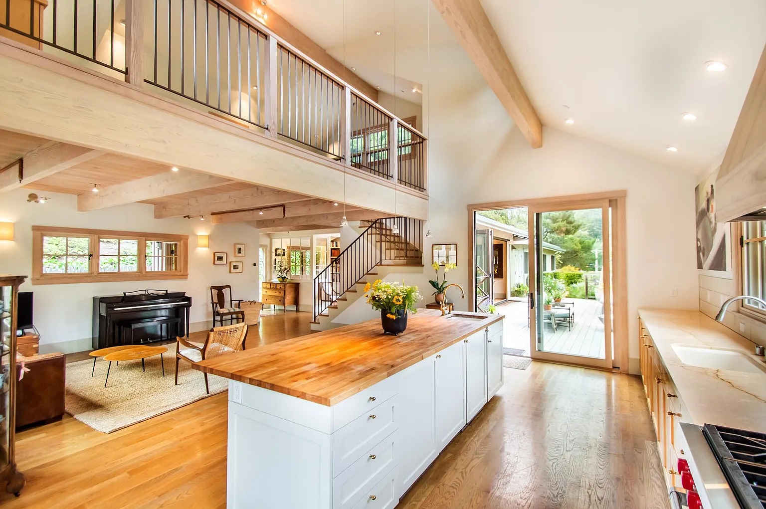 Open-concept living room and kitchen with wooden beams, hardwood floors, large sliding glass door leading to outdoor deck, kitchen island with flowers, and cozy seating area.