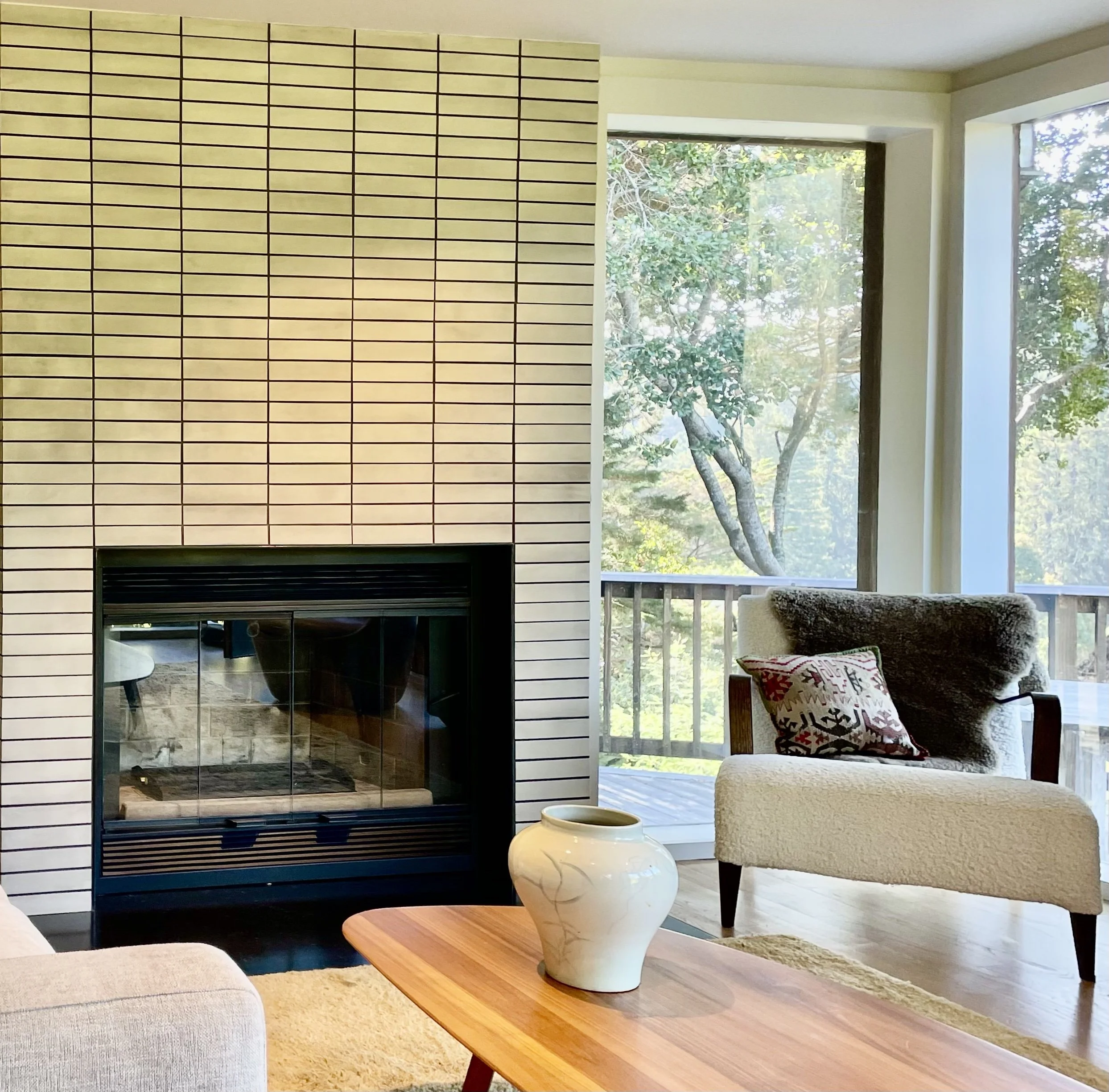 Living room with a fireplace, a side chair with a blanket and a patterned pillow, a wooden coffee table with a ceramic vase, large windows with a view of trees outside.