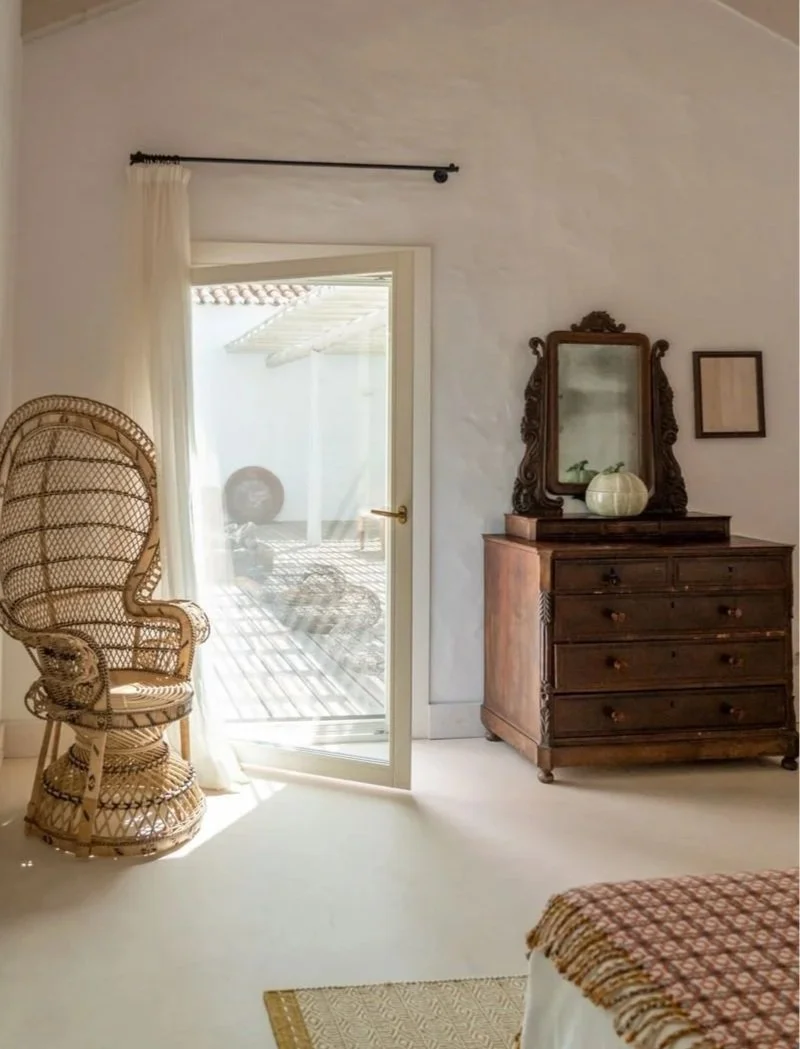 Sunlit bedroom with wicker chair, wooden dresser with mirror, and glass door leading to outdoor patio.