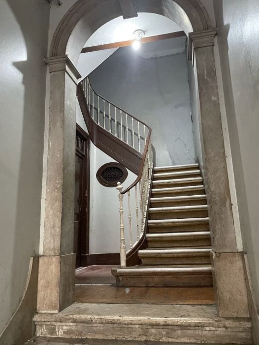 A curved wooden staircase with a white railing ascending to a second floor, viewed through an arched doorway with stone columns. The wall on the right side shows signs of wear, and a ceiling light illuminates the area.