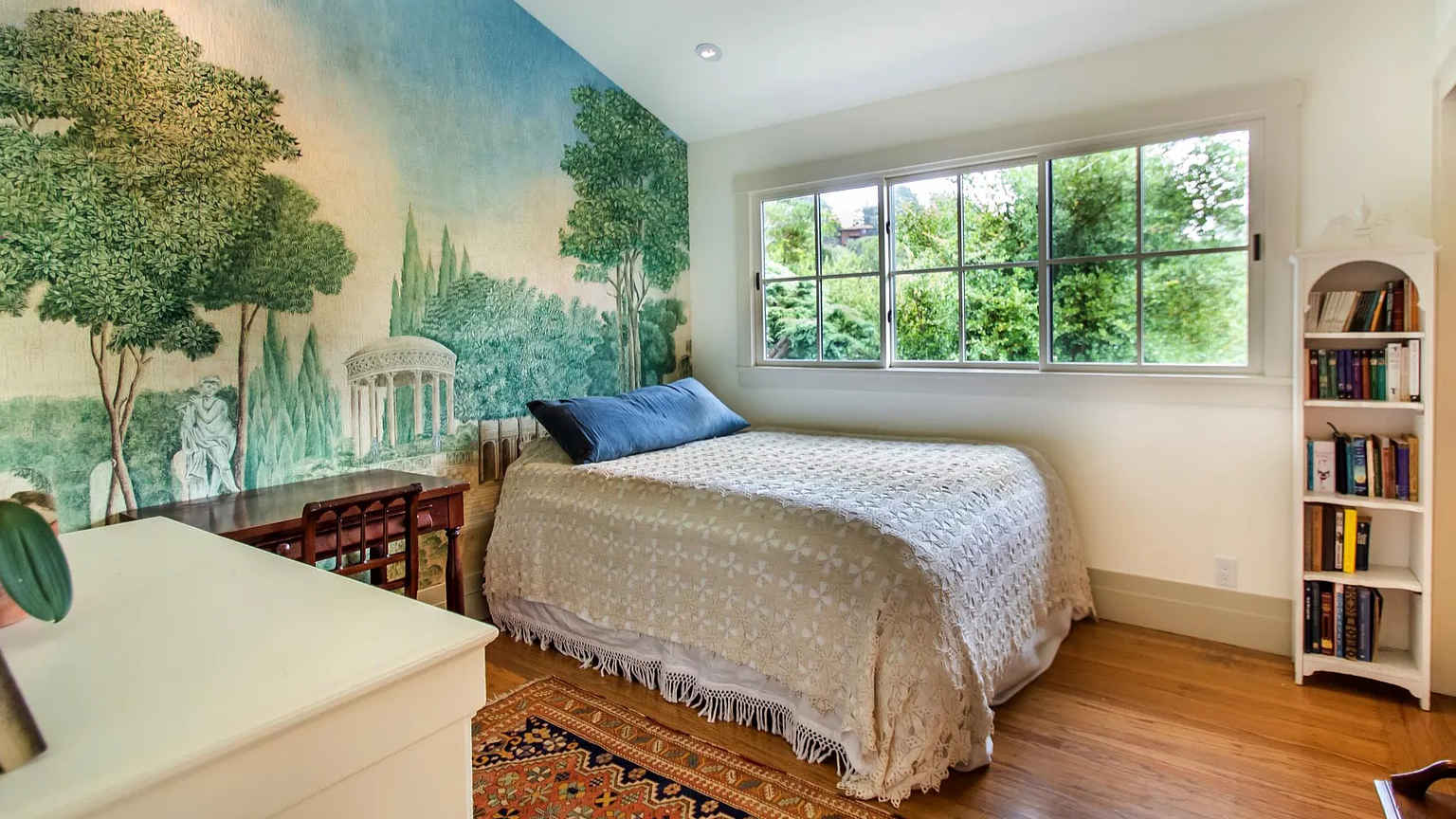 A bedroom with a large window showing greenery outside, a bed with a white textured bedspread and a dark blue pillow, a white bookshelf filled with books, a wooden table, and a large mural of trees and ancient ruins on the wall.