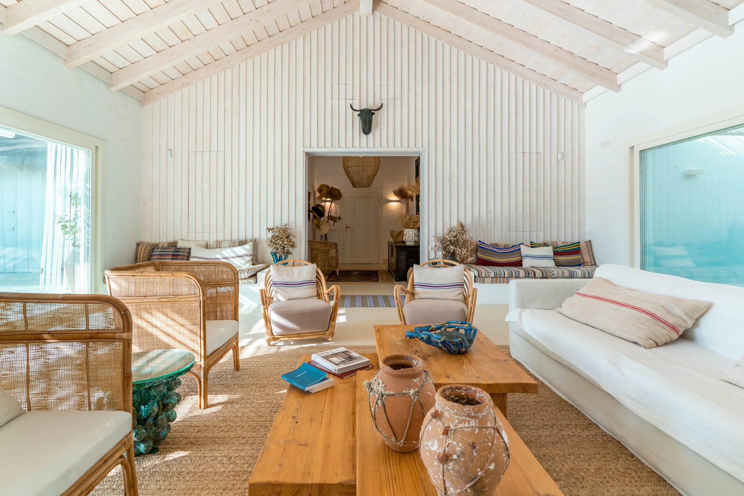 Living room with white walls, high ceiling with exposed wood beams, large glass sliding doors, rattan chairs, white sofa with striped pillows, wooden coffee table, and decorative vases, with a hallway in the background.