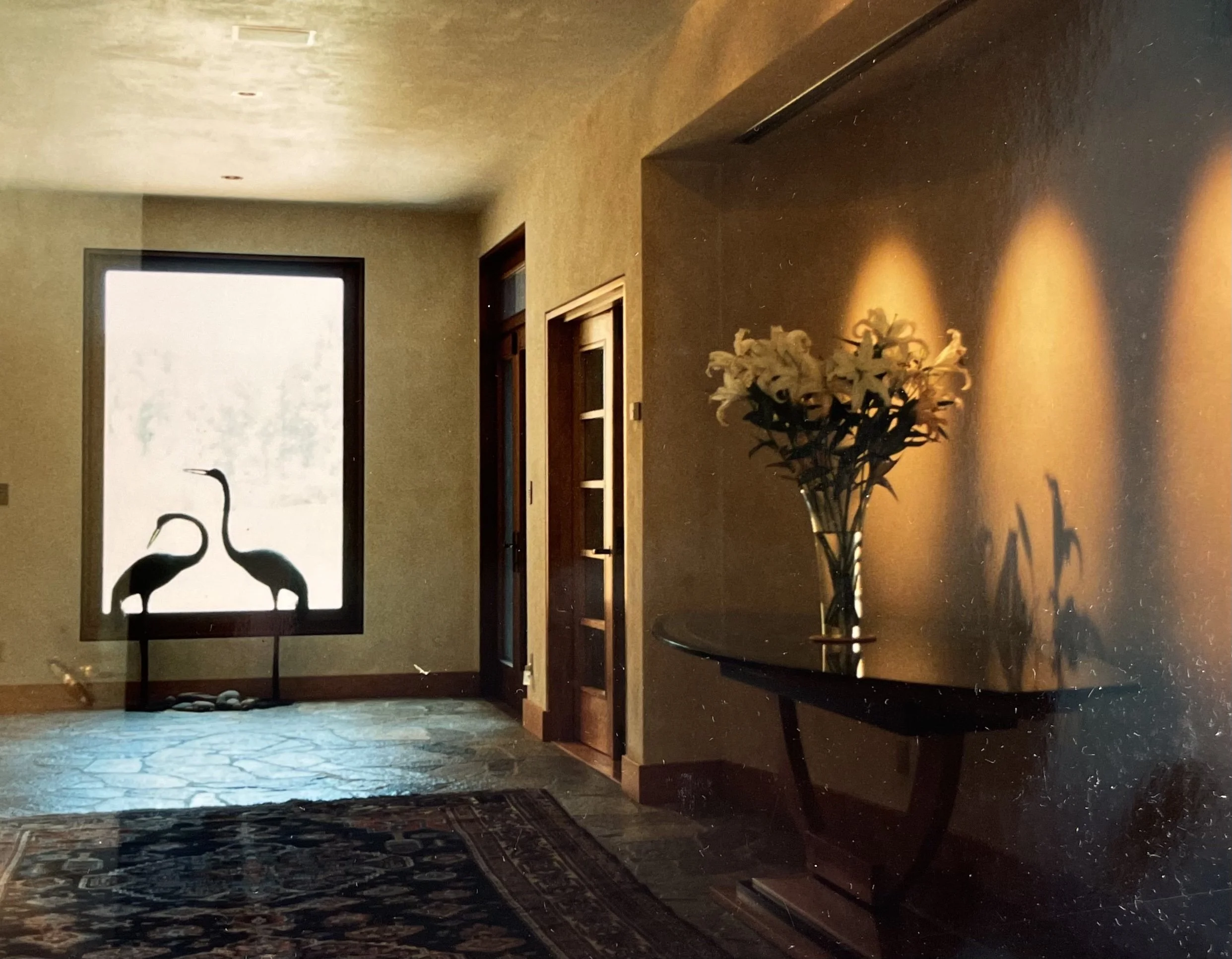 Interior room with a window featuring two flamingo silhouettes, a side table with a vase of white lilies, and warm lighting casting shadows on the wall.