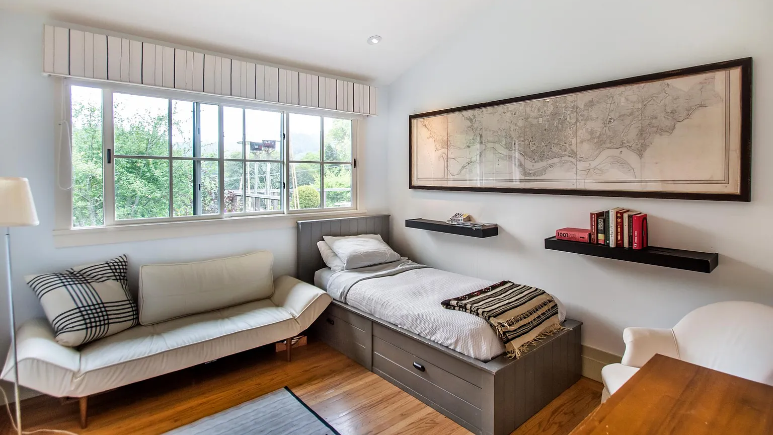 A cozy bedroom with a window view of green trees. It contains a single bed with a gray headboard, a white sofa with patterned pillows, a white armchair, and a wooden table. Two floating shelves hold books and a remote control. A large framed map decorates the wall above the bed.