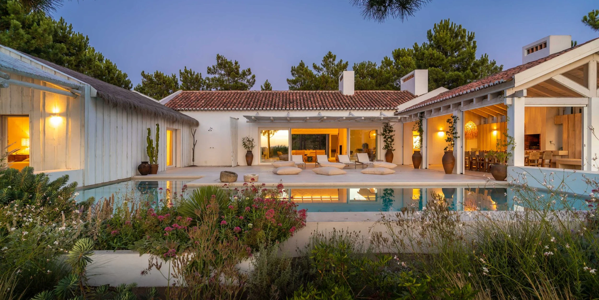 A modern house with white walls and a terracotta-tiled roof, featuring a pool in front surrounded by plants, lounge chairs, and large planters, with interior lights glowing through open doors and windows at dusk.