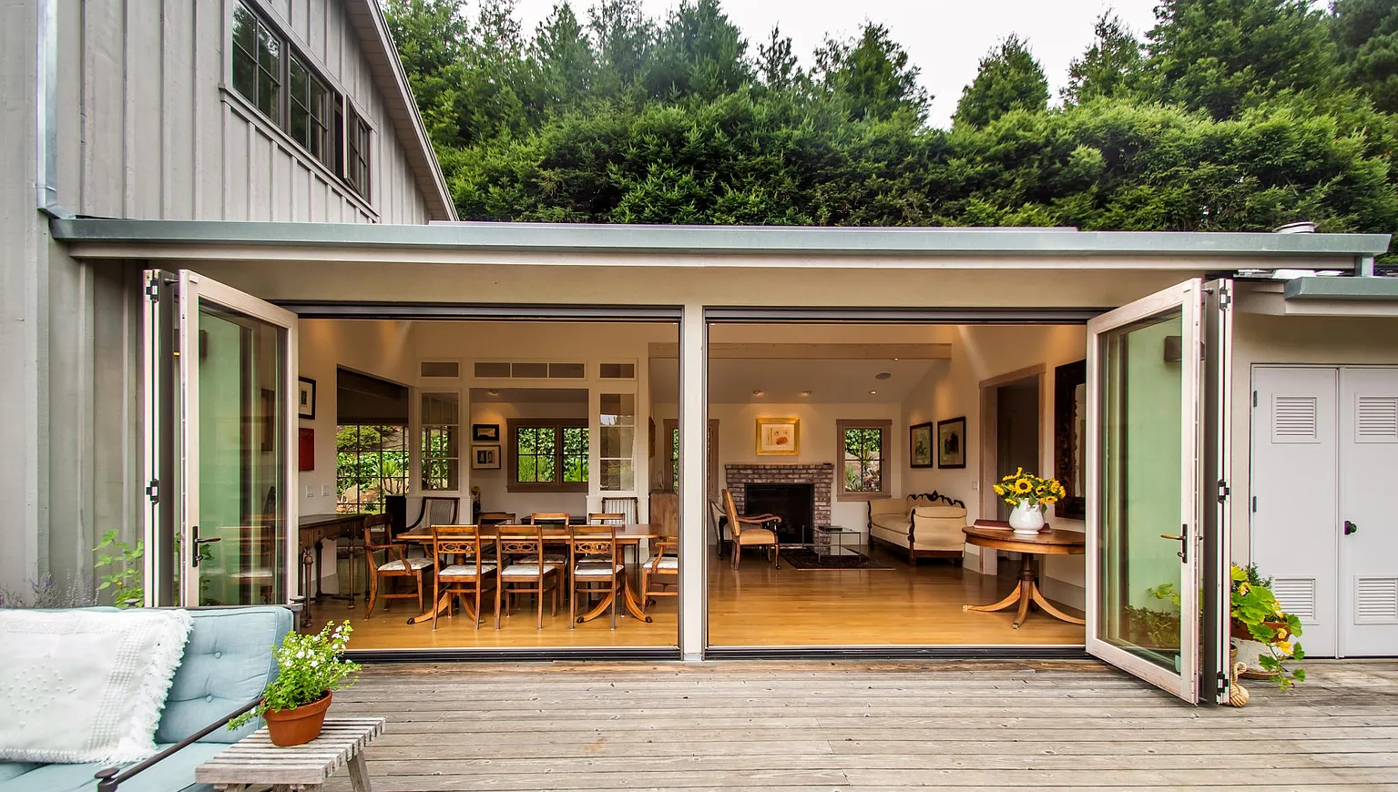 View of a house with large open French doors leading to a wooden deck, revealing an interior with a dining table, chairs, and a living room with a fireplace, surrounded by green trees.