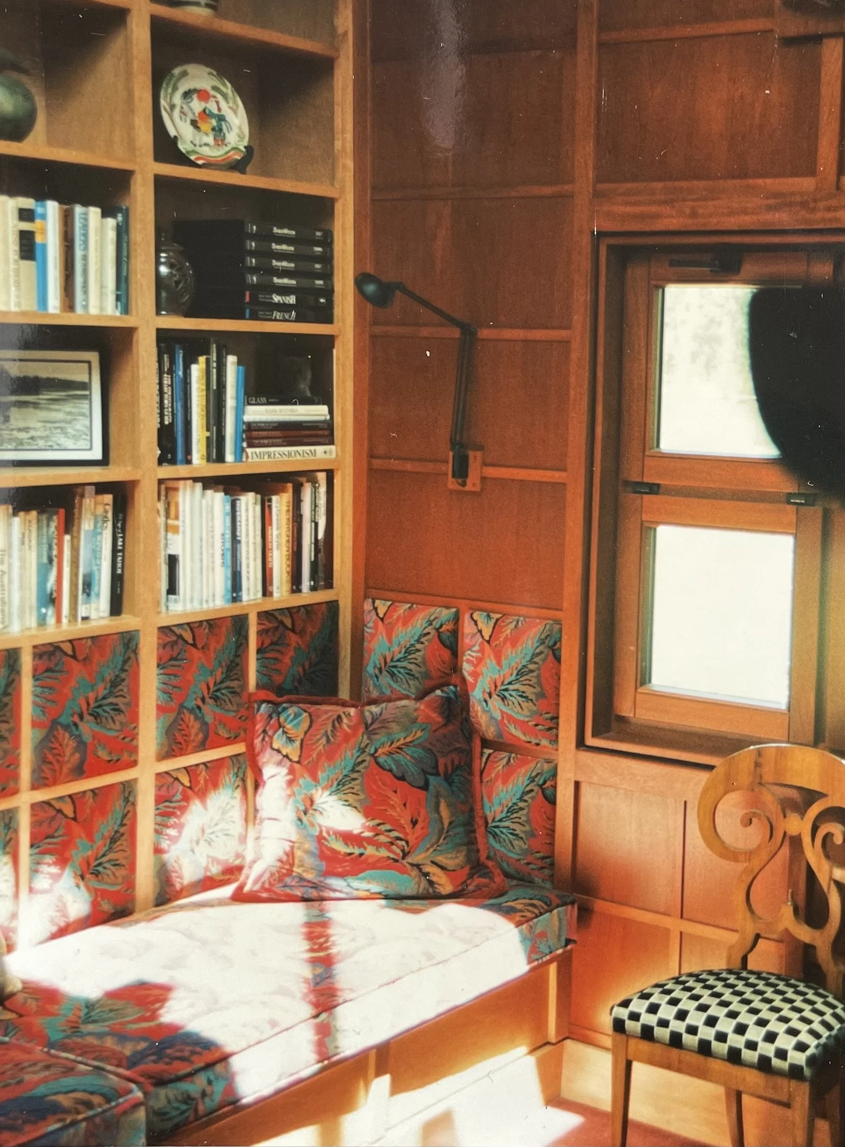 A cozy reading nook with a built-in wooden bookshelf filled with books, decorative items, and a black stereo system. Next to it is a cushioned bench with a colorful, leafy pattern pillow, and a wooden chair with a black-and-white checkered seat. The room has warm wooden paneling, two small windows, and a wall-mounted adjustable reading lamp.