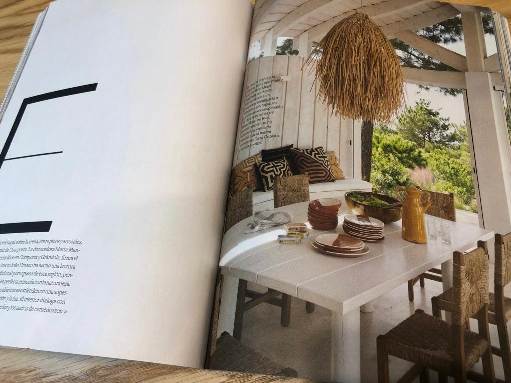 Indoor dining area with a white table set with dishes and a yellow pitcher, surrounded by woven chairs, with a large straw light fixture hanging from the ceiling and a lush green view outside.