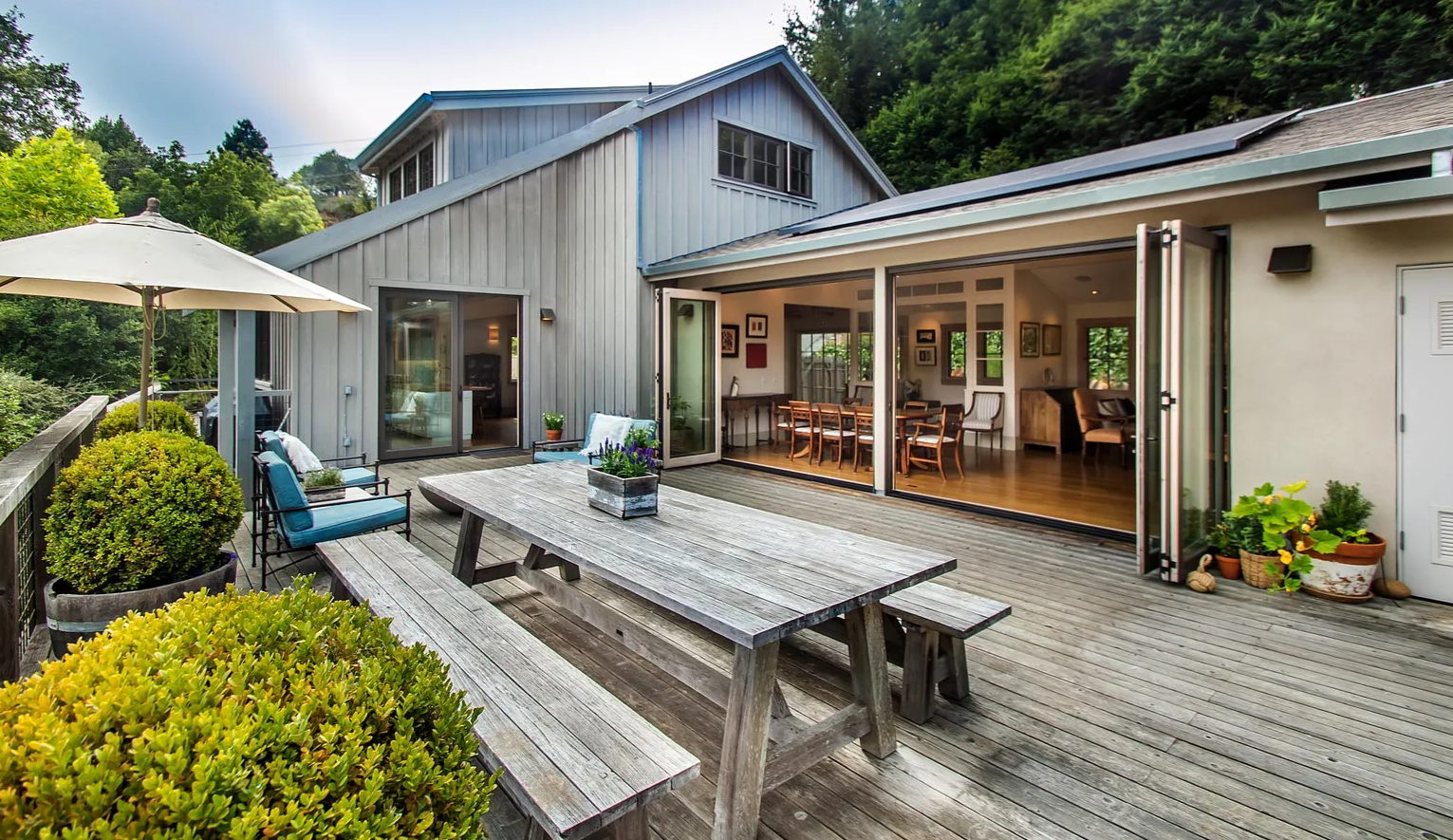 Exterior view of a wooden deck attached to a house with large glass doors opening to a dining and living area, outdoor seating with a table and bench, potted plants, and surrounding greenery and trees.