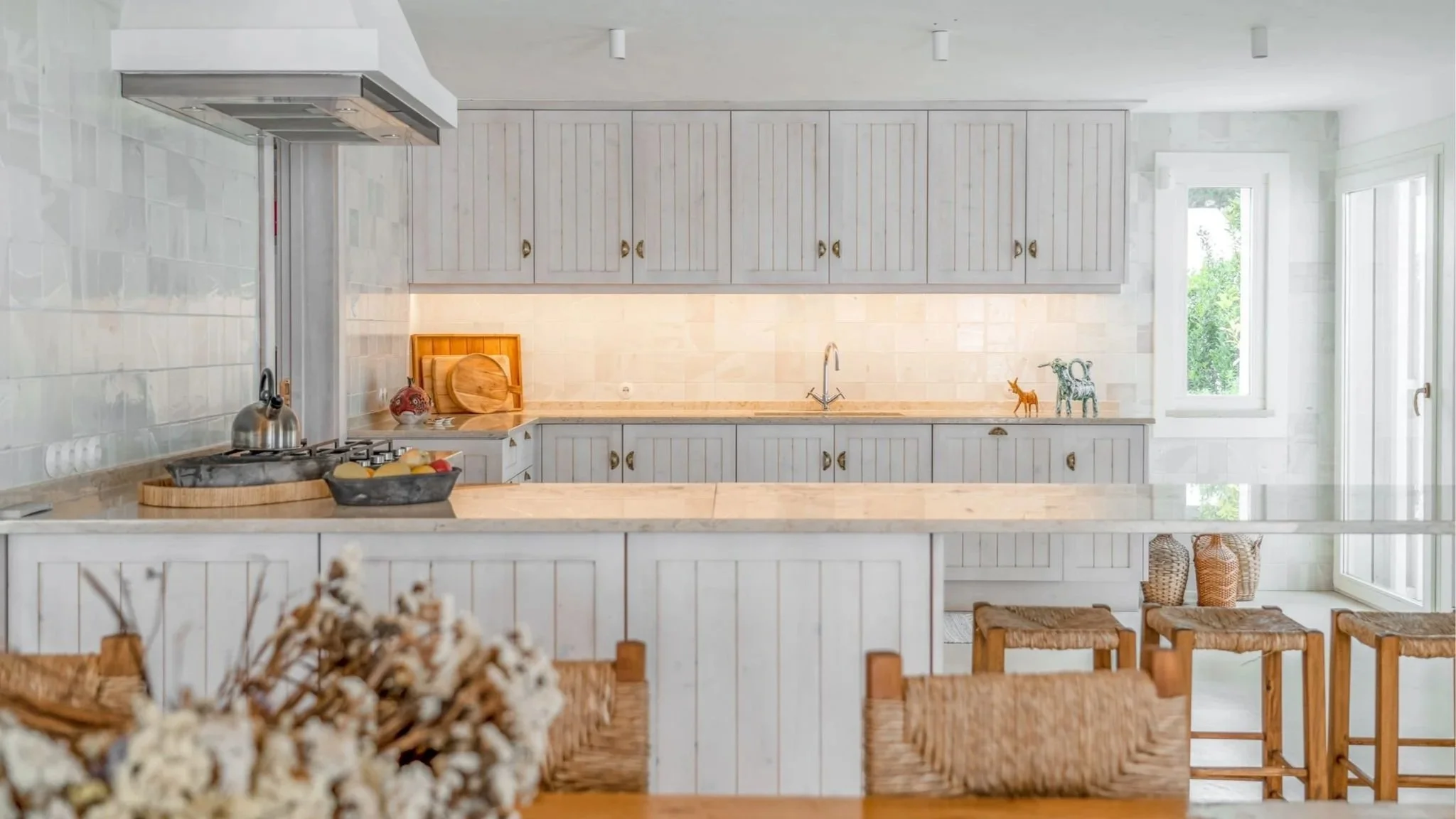 Bright, airy kitchen with white wood cabinets, beige tiled backsplash, and large windows letting in natural light. The countertop has a wooden cutting board, a bowl of fruit, and a teapot. There are wicker baskets and wooden stools at the kitchen island.