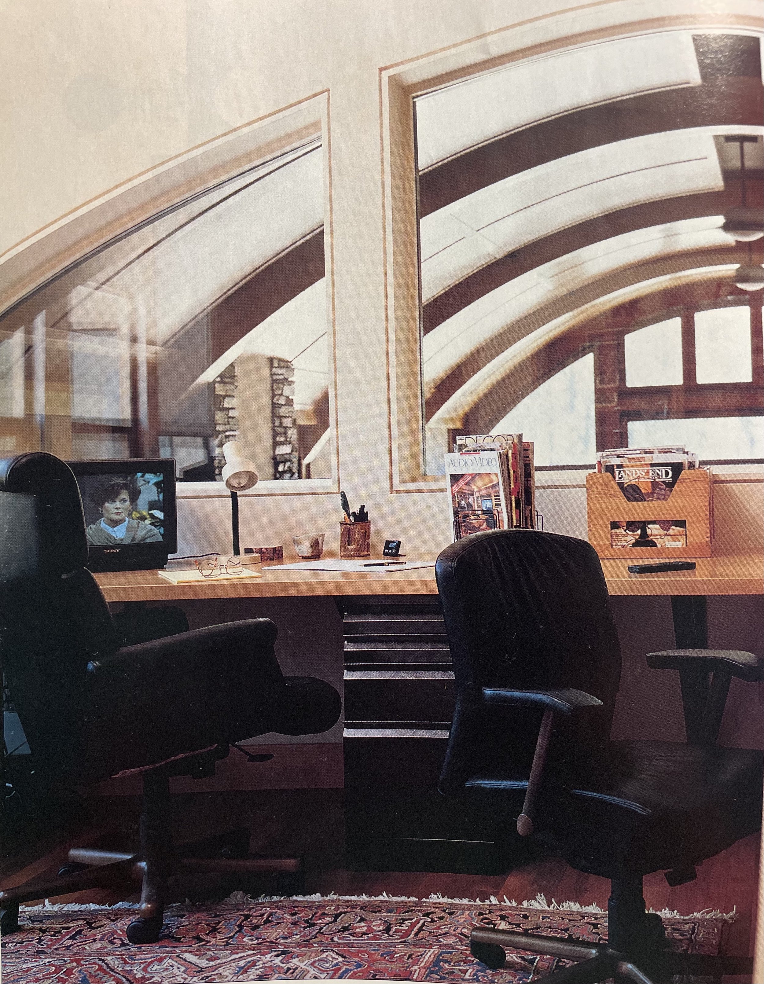 An office with a wooden desk, two black office chairs, magazines on a wooden holder, a small television showing a woman's face, a desk lamp, pens, and glasses, with large arched windows revealing a curved building exterior.
