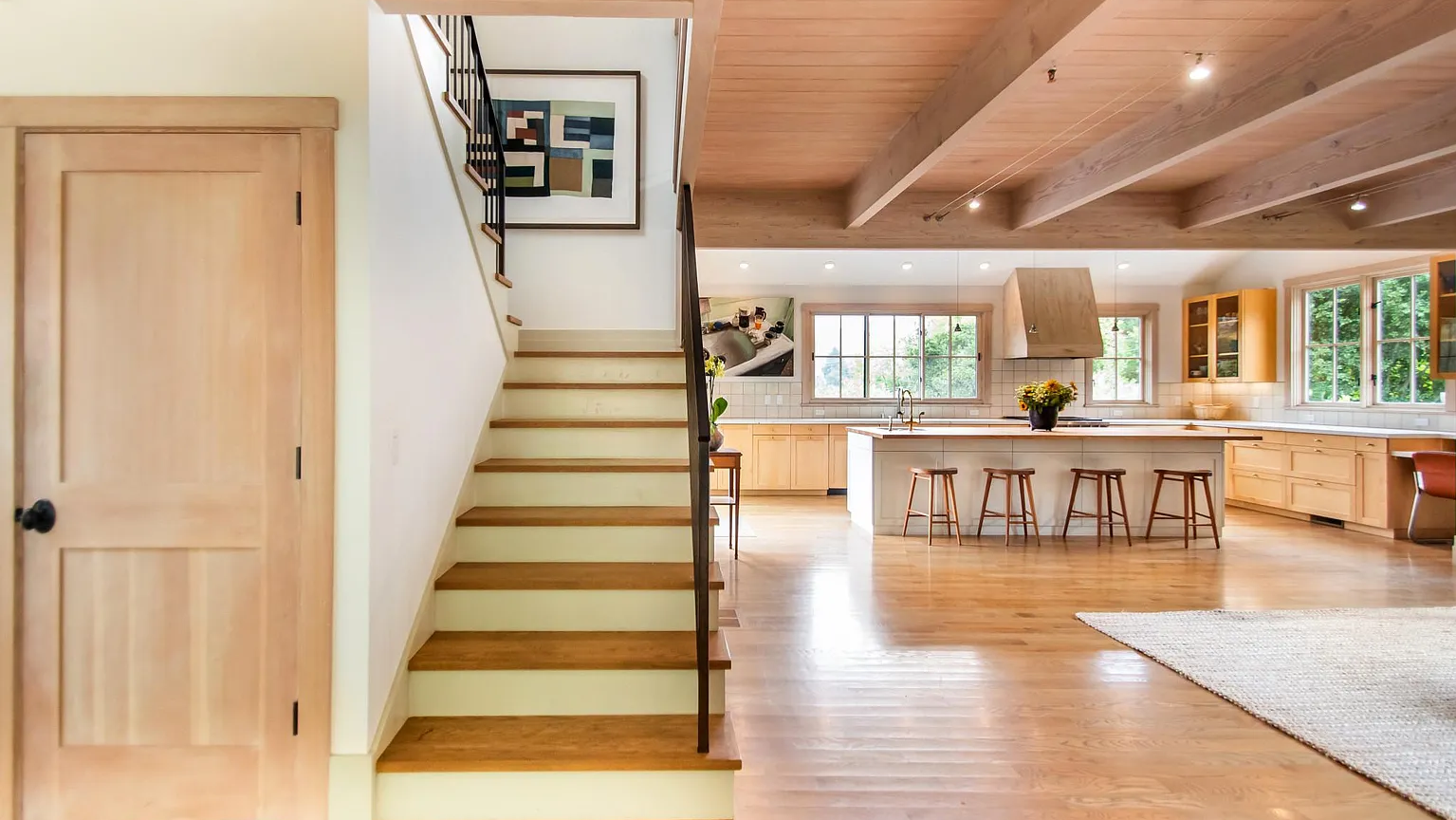 Open-concept kitchen and living area with wooden floors, a kitchen island with barstools, large windows, wooden ceiling beams, and minimal decor.