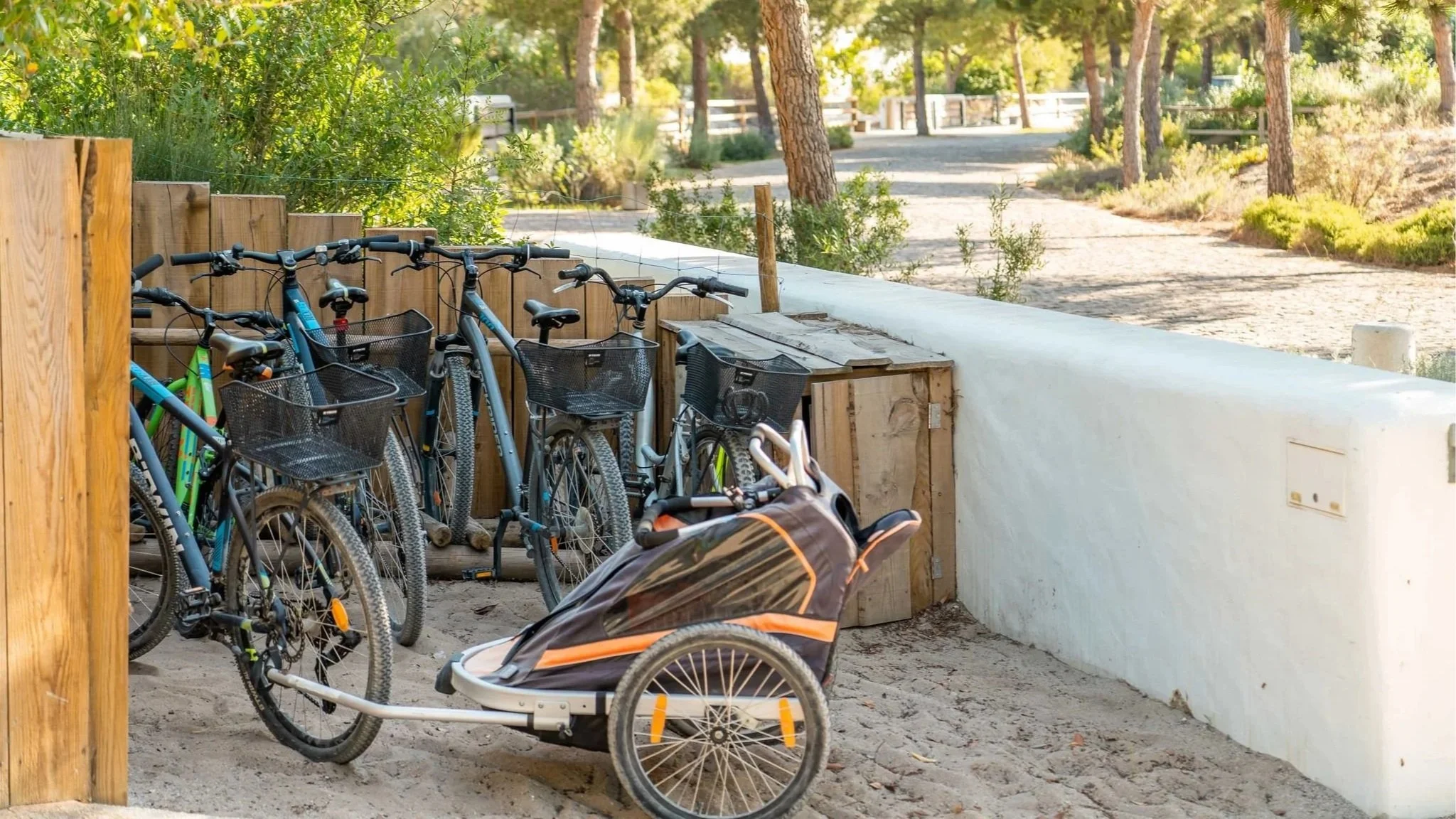 Several bicycles and a child trailer parked near a wooden fence in a sandy outdoor area with trees and a dirt trail.