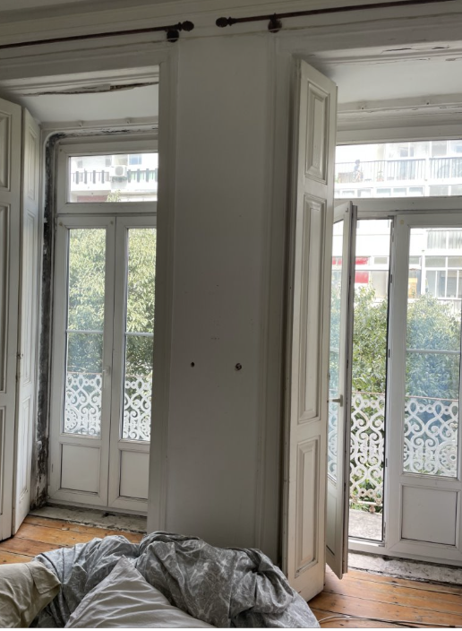 Interior view of a room with two large windows and a door leading to a balcony, partially open, with white window frames and shutters, revealing outdoor trees and buildings.