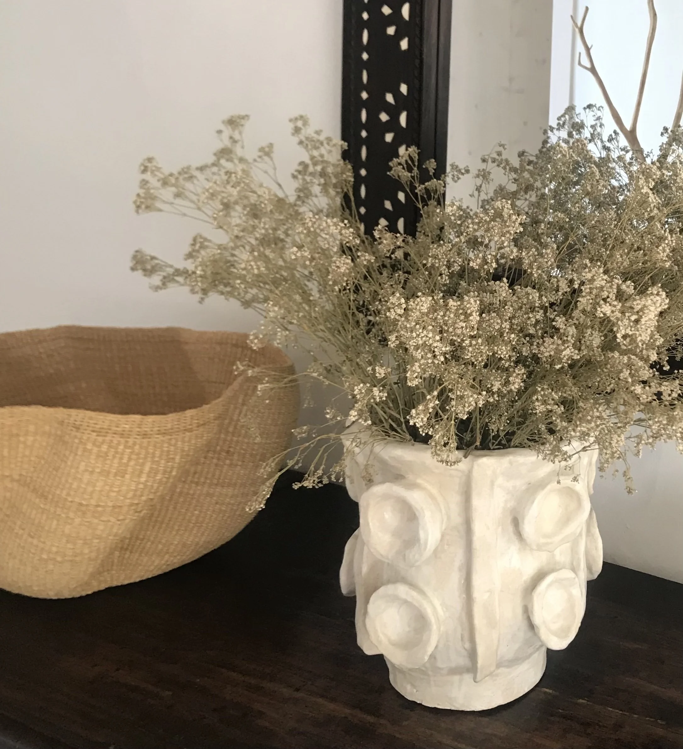 Decorative white ceramic vase shaped like a face with circular features and a pointed nose, holding dried white flowers, on a dark wooden surface, with a woven basket nearby.